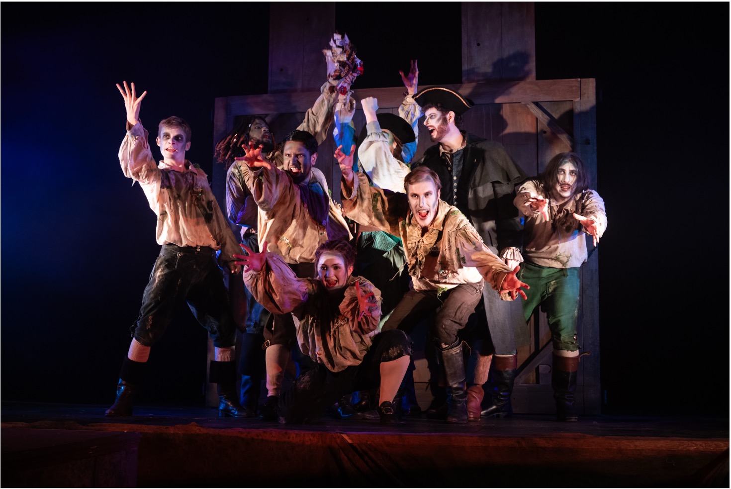A group of eight performers in period costumes pose dramatically on stage. They are dressed in ragged clothing, with some showing face paint or makeup, against a backdrop of wooden panels and low lighting, creating a theatrical atmosphere.