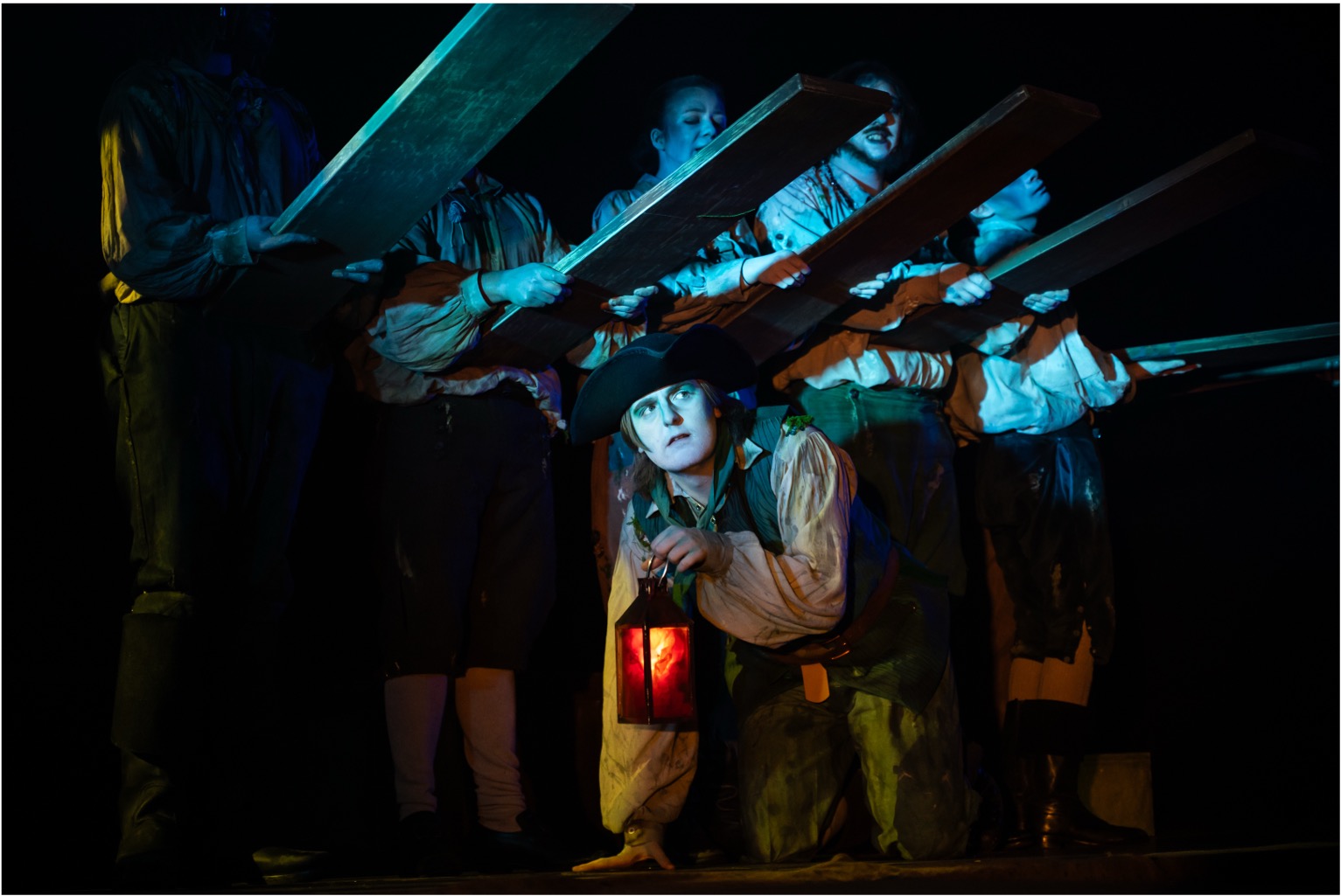 A group of performers dressed in historical costumes stands in a dimly lit setting, holding long wooden planks above their heads. One performer crouches in front, holding a lantern and looking towards the audience, while others are positioned behind, slightly obscured by the planks.