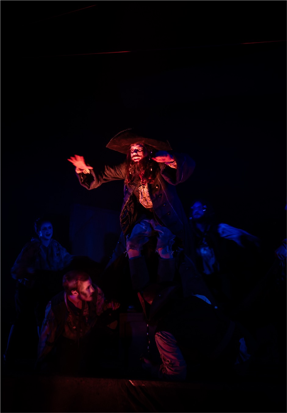 A performer dressed as a pirate, wearing a hat and dark coat, stands on a stage, gesturing dramatically. Surrounding figures in period costumes are partially visible in low light, creating a theatrical scene.