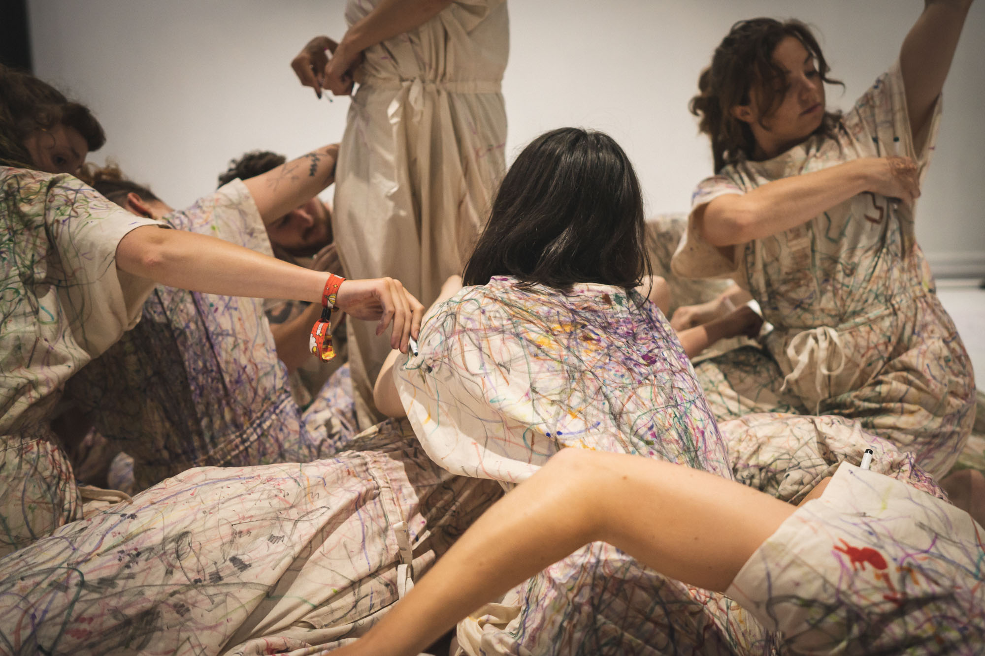 Several people in loose, patterned dresses are seated and reclining closely together in a gallery-like room with white walls.