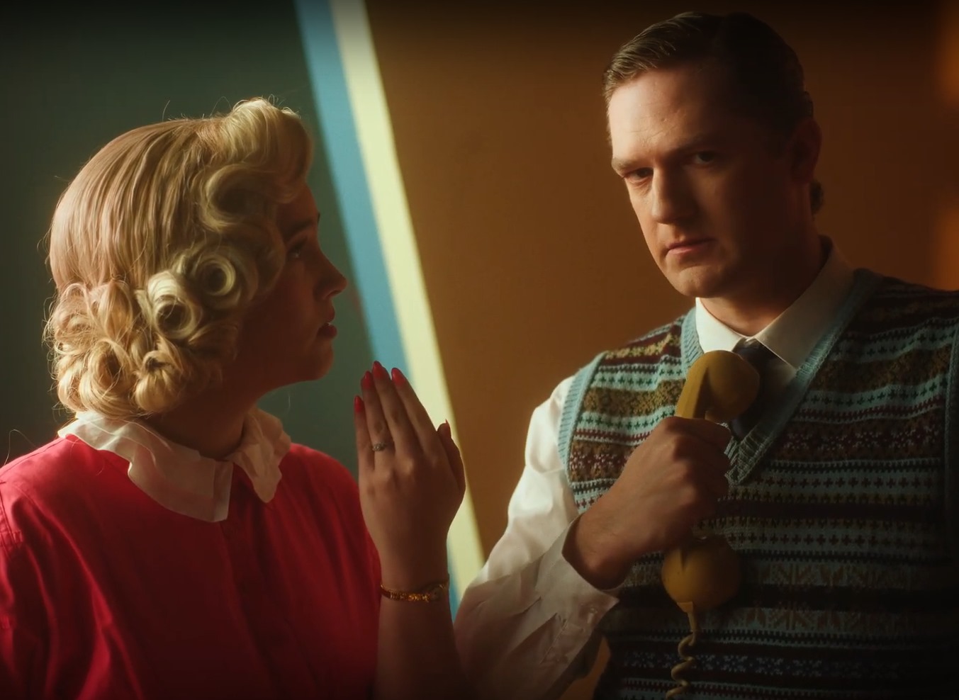Blonde woman in red cardigan and brunette man in patterned vest face each other, touching hands with blue lightsaber between them.