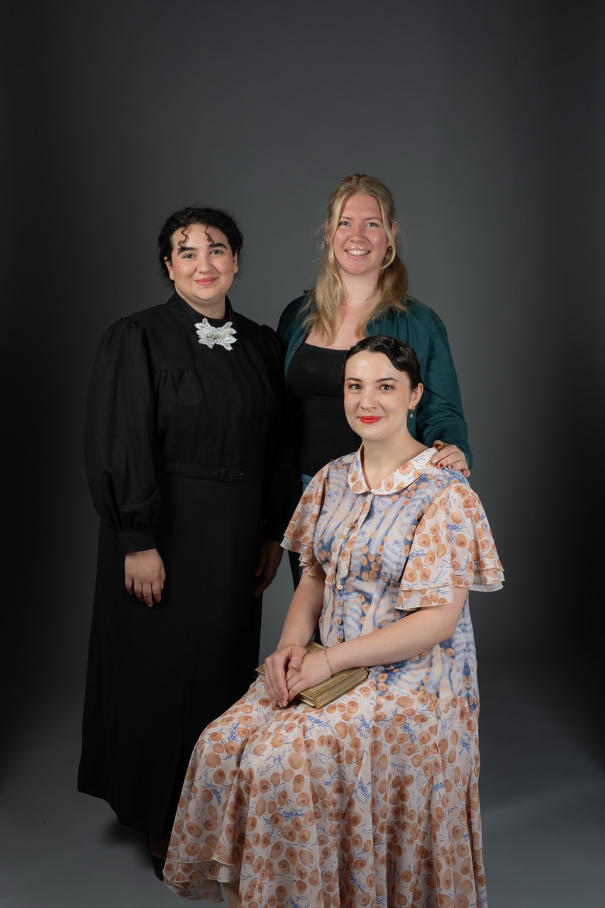 Three women posing together against a grey background. The woman on the left wears a black dress with a white collar, the woman in the middle has long blonde hair and wears a dark green top, and the woman on the right is seated in a floral dress with a handbag on her lap.