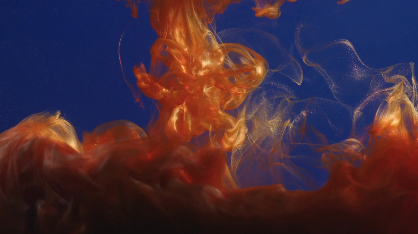 An abstract image showing swirls of orange and gold liquid. The vibrant colours blend with a dark blue background, creating a dynamic visual effect. The liquid appears to be flowing from the top, forming intricate patterns as it disperses.