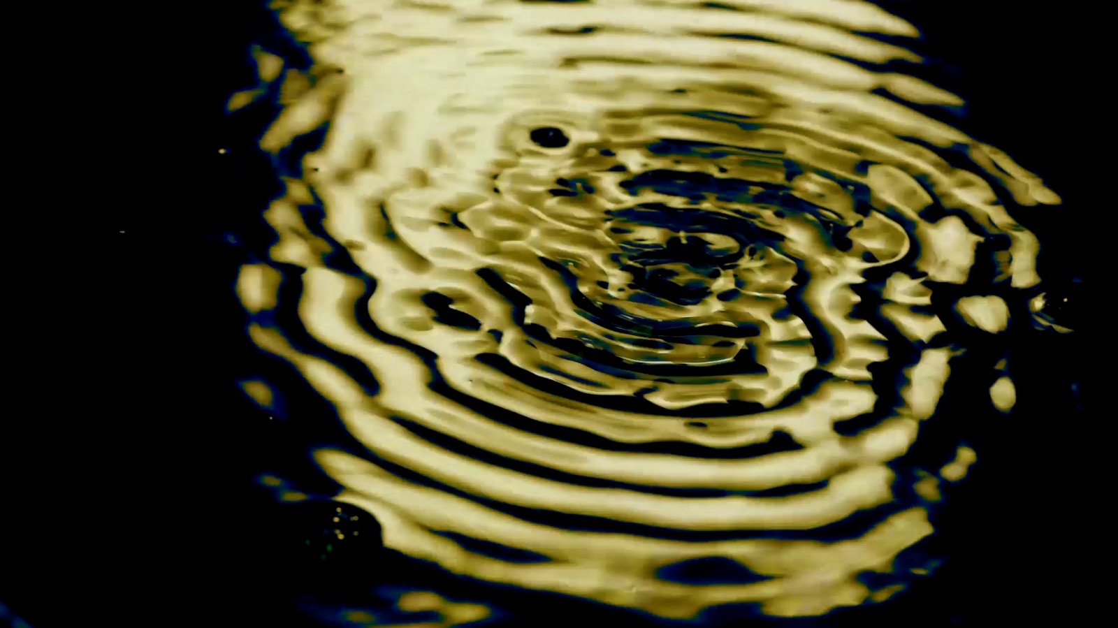 Close-up of water ripples reflecting golden light, creating a series of concentric rings on the surface. The background is dark, emphasizing the luminosity of the water and the pattern of the ripples.