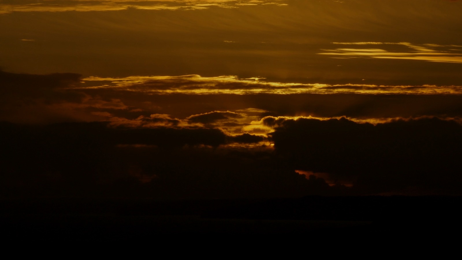 A twilight sky filled with dark clouds illuminated by a golden sunset. The horizon features a range of cloud formations with varying shades of orange and yellow.