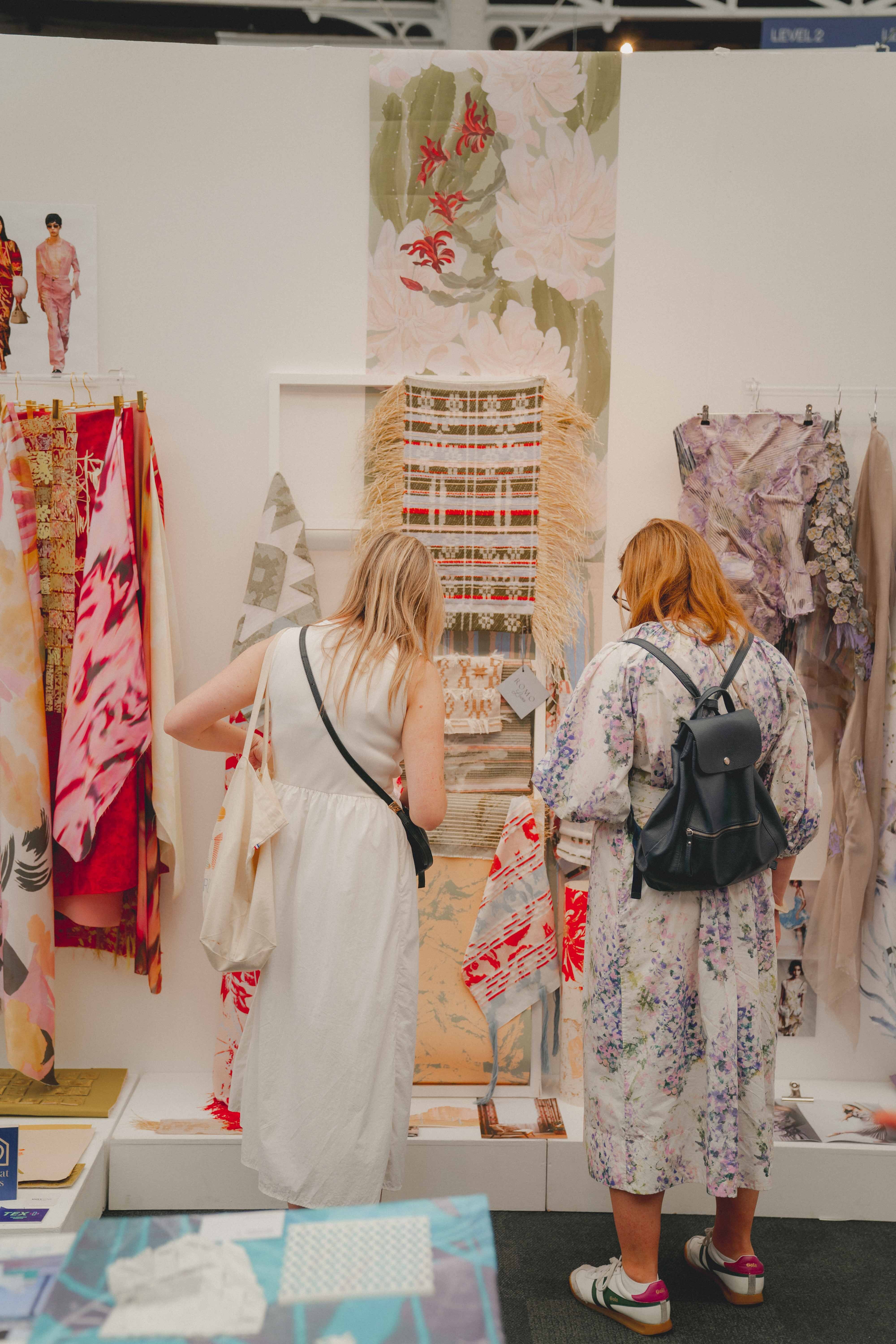 Two people viewing textile display at trade show booth with floral fabrics, pattern samples, and design boards on white walls.