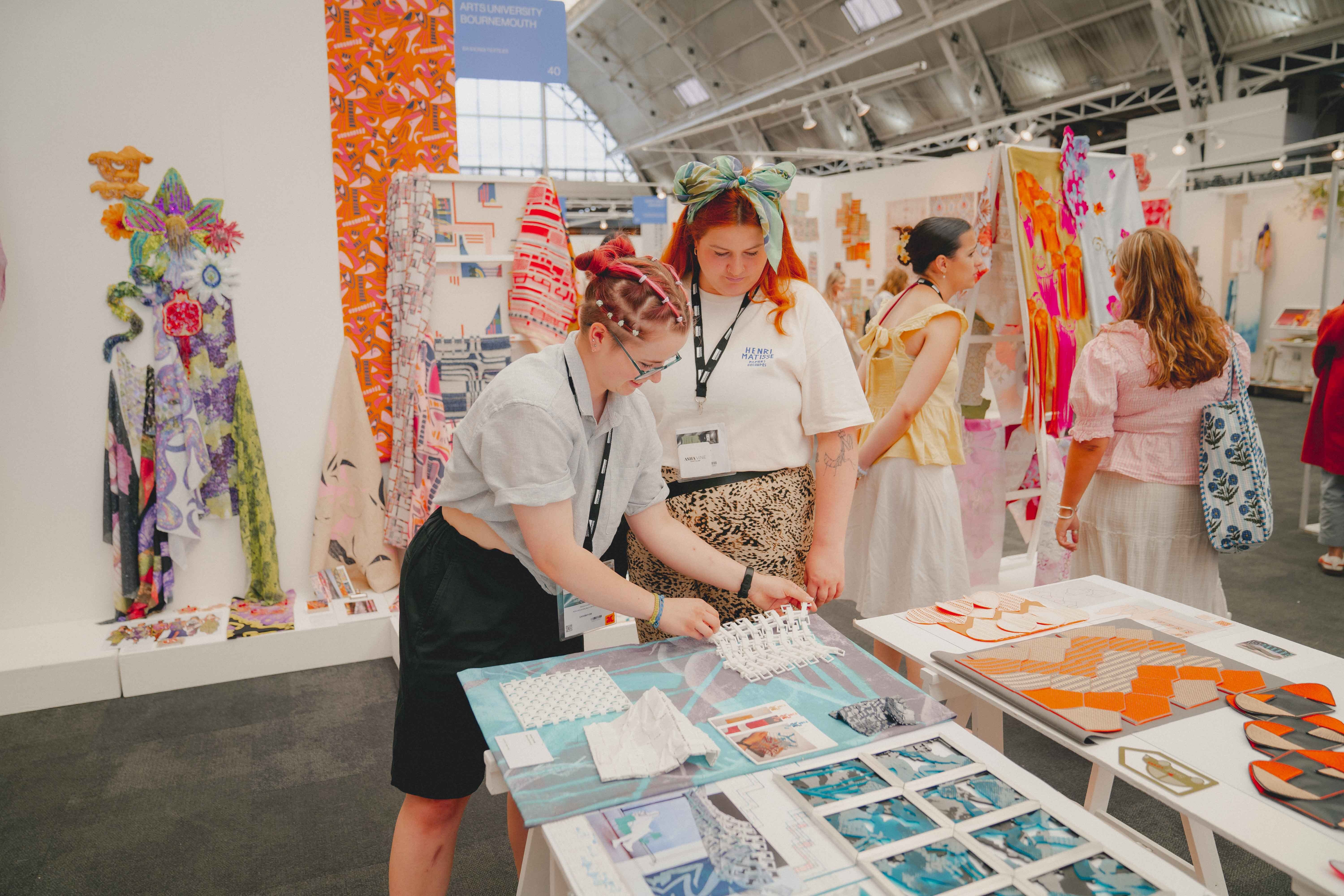 Two people examining fabric samples at white table in exhibition space with colourful textile displays and industrial ceiling.
