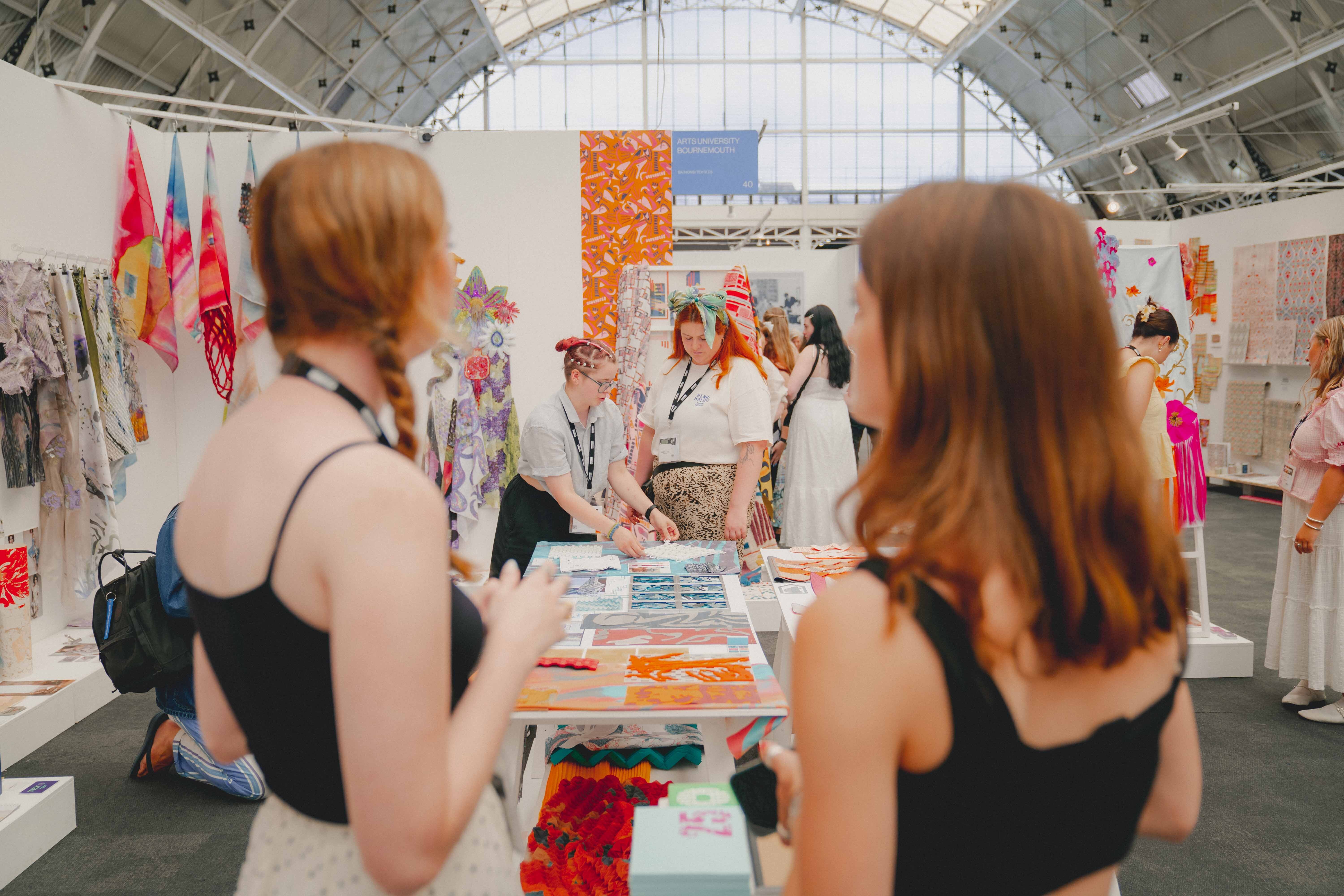 Two women with blonde hair at white table in exhibition hall with glass ceiling, colourful artworks and banners displayed on walls.