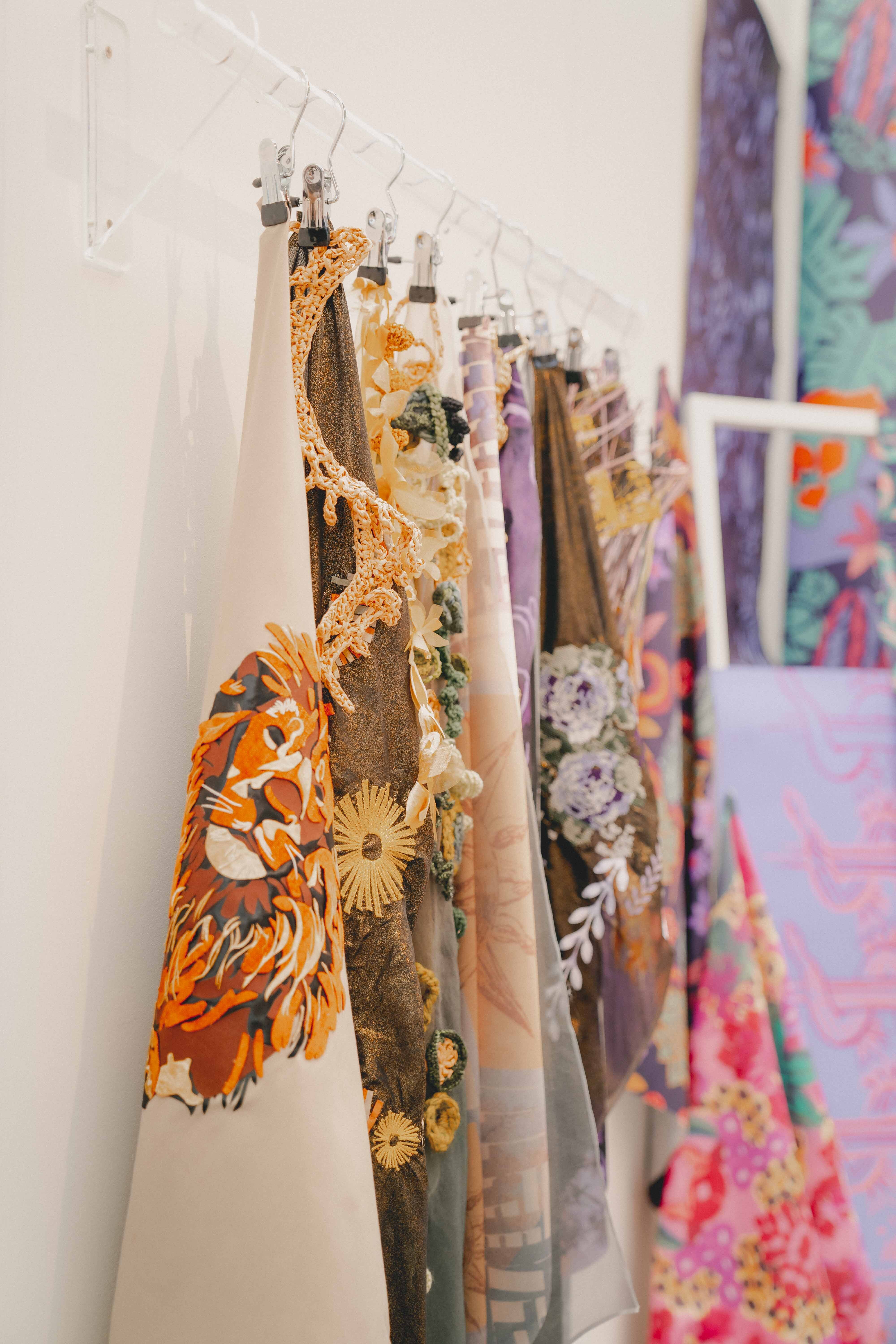 Colourful fabric samples hanging on clips from horizontal rail against white wall, featuring floral and abstract patterns in orange, green, purple, and pink tones.