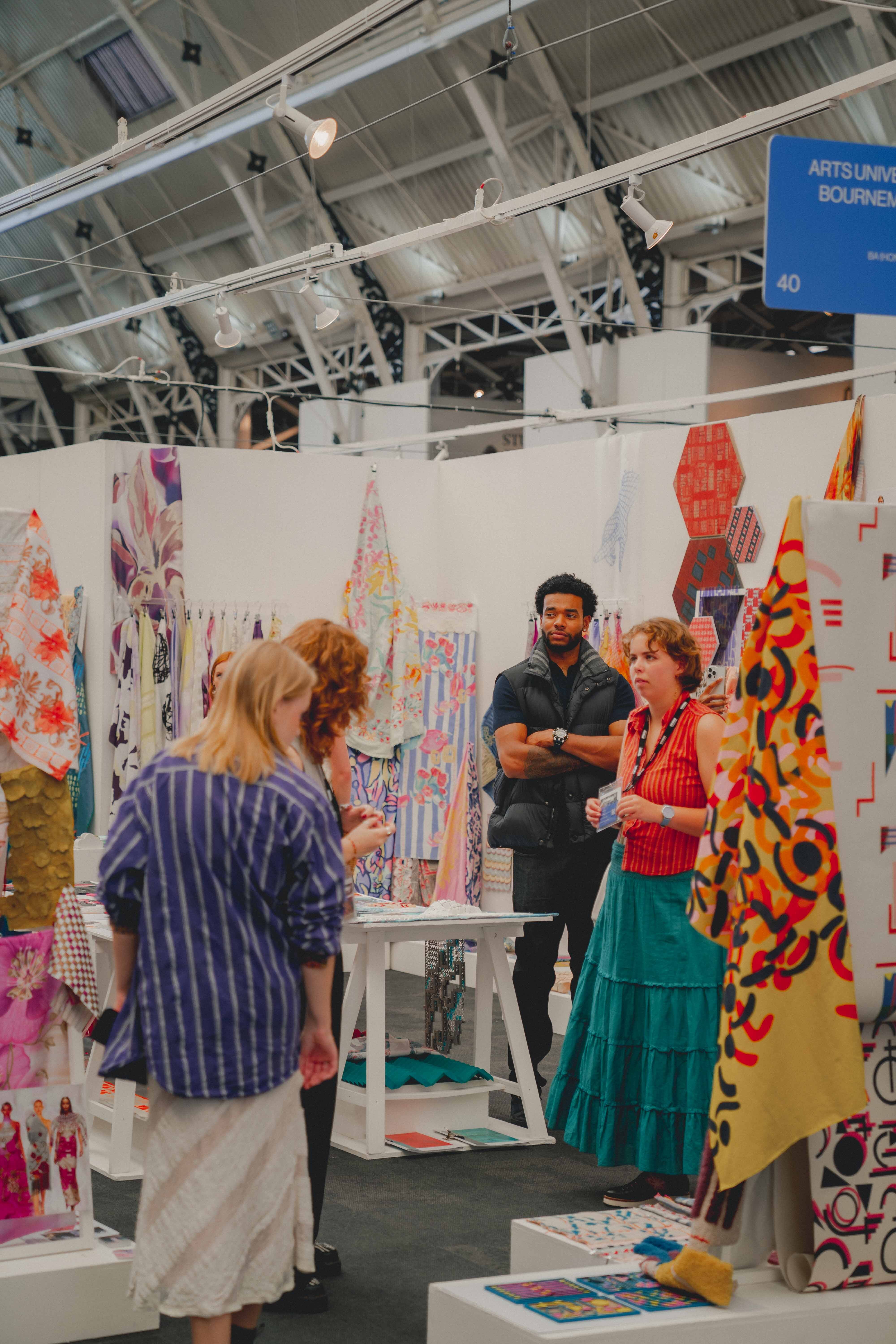 Textiles exhibition space with white booth displaying colourful textile works. Visitors browse fabric pieces in yellow, red, and green patterns.