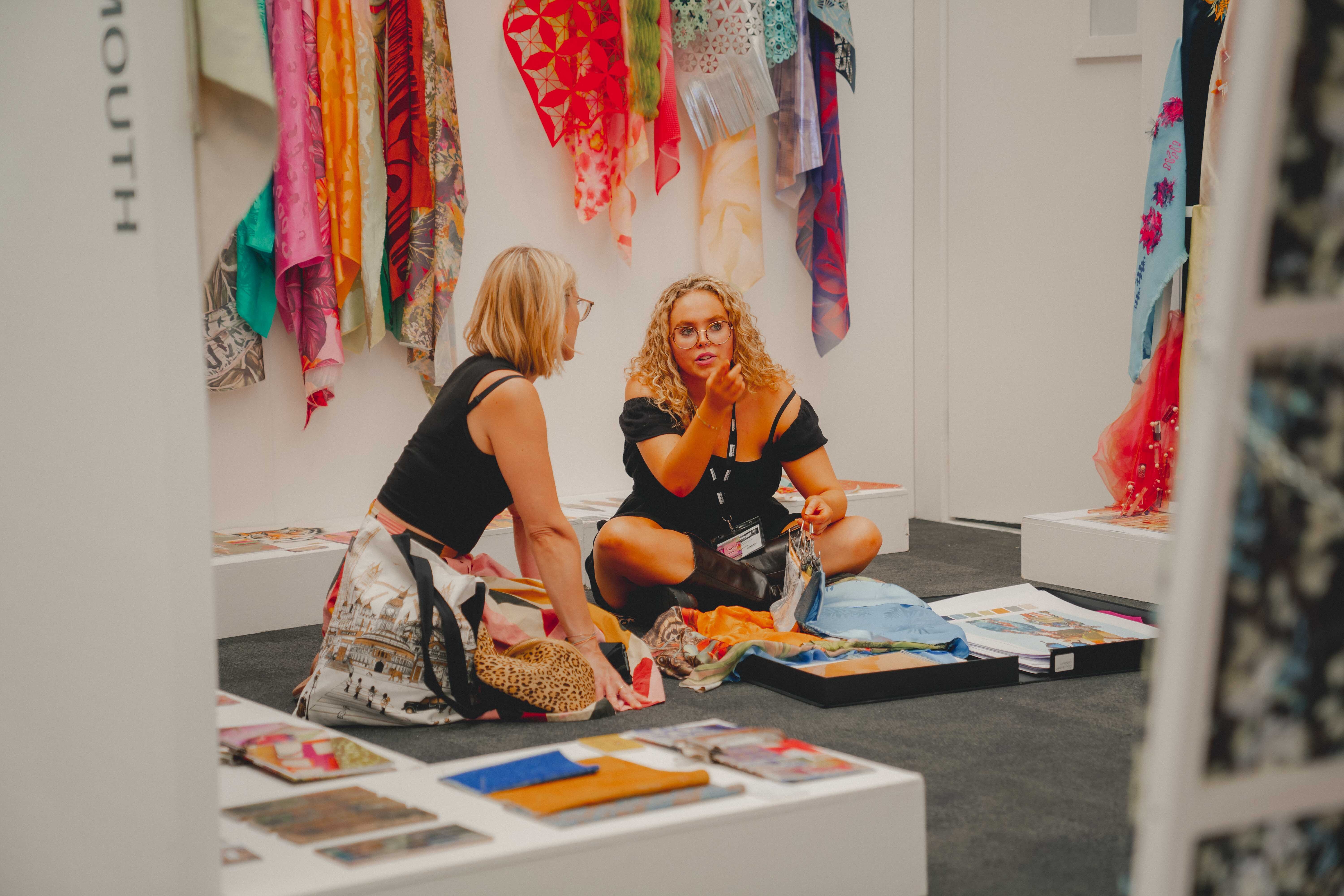 Two people sitting on gallery floor surrounded by colourful fabric samples and art materials, with vibrant textiles hanging above.