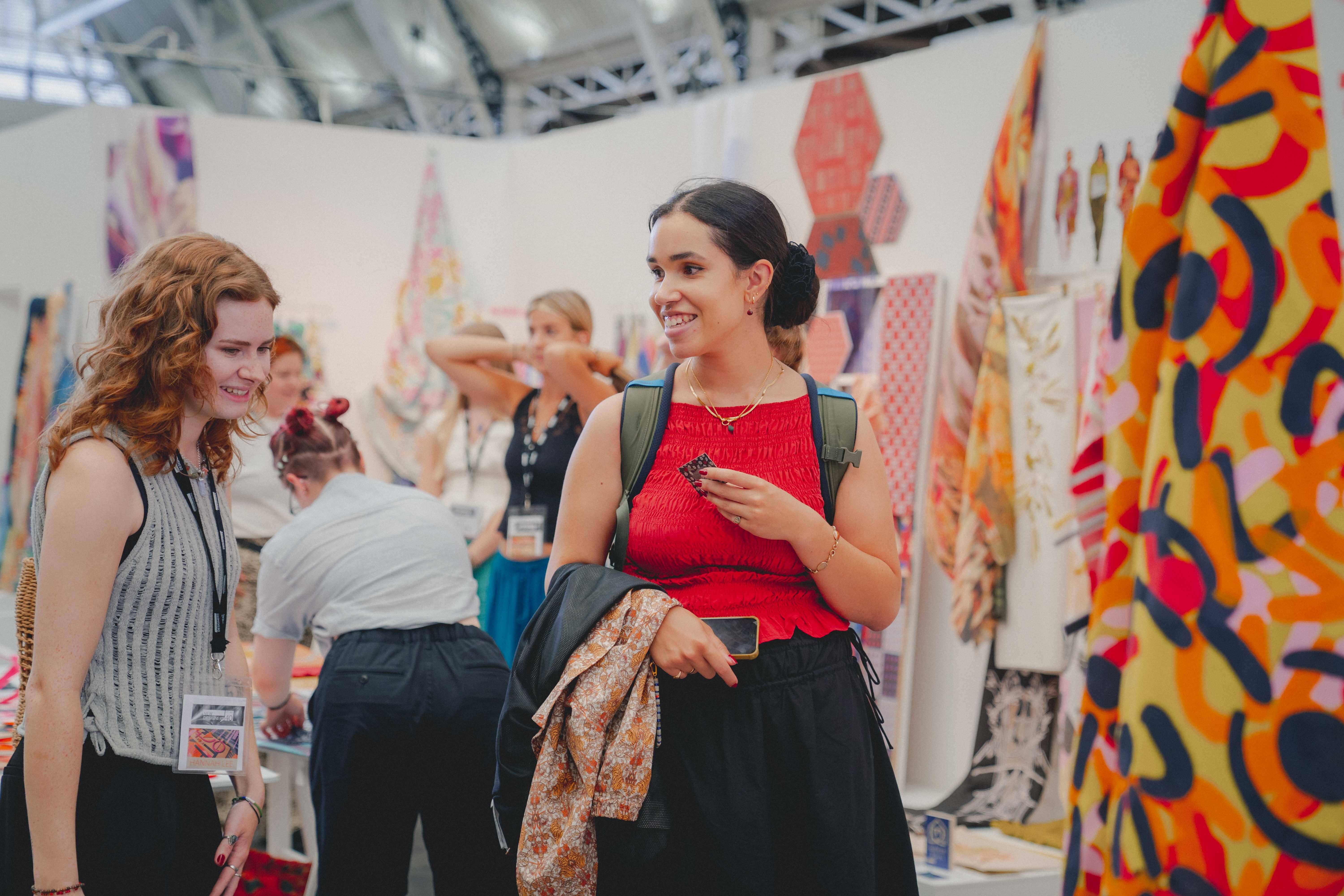 Two women chatting at textiles exhibition with colourful abstract paintings displayed on white walls in background.