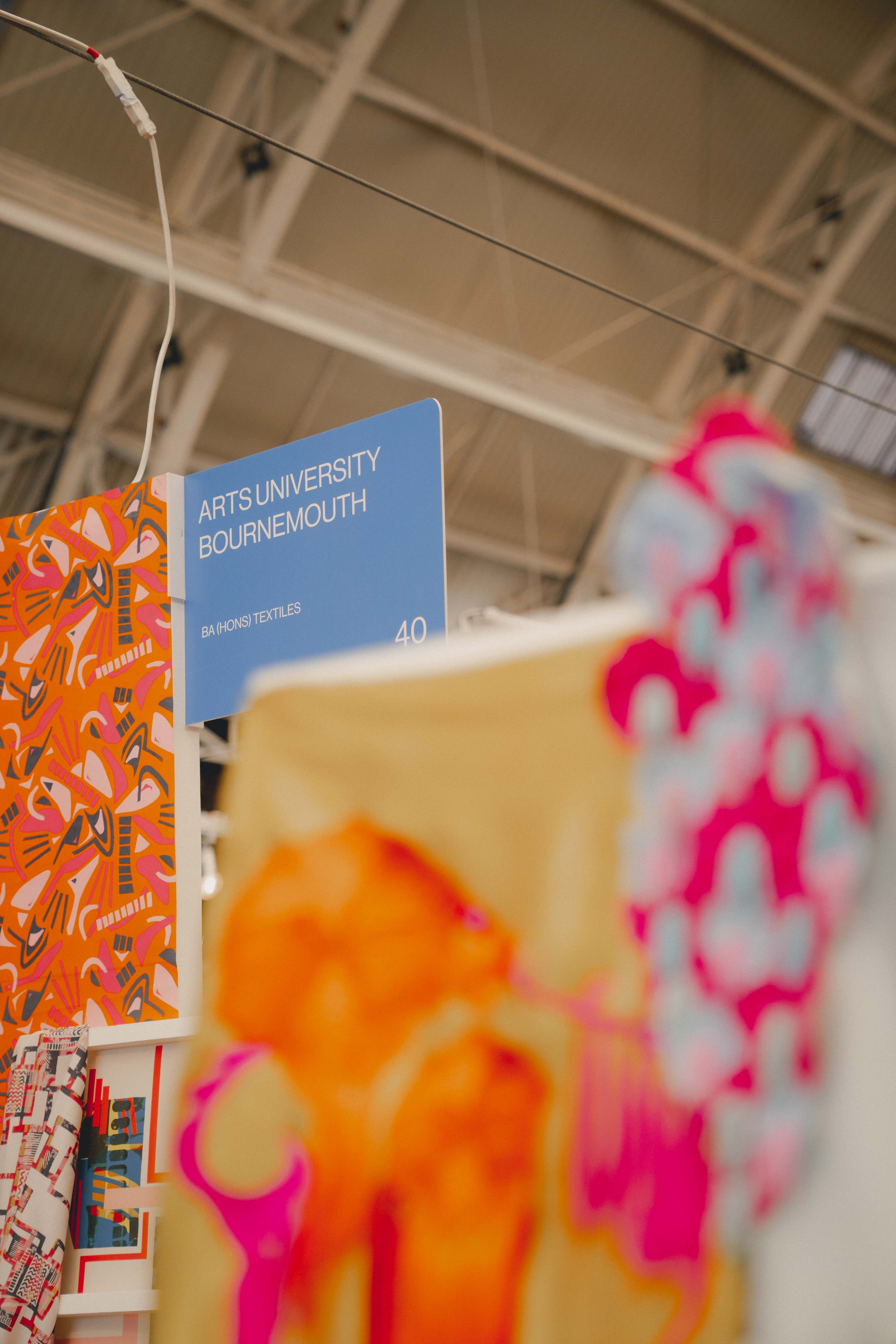 Blue Arts University Bournemouth sign suspended above blurred orange and pink fabric or clothing display in warehouse setting.