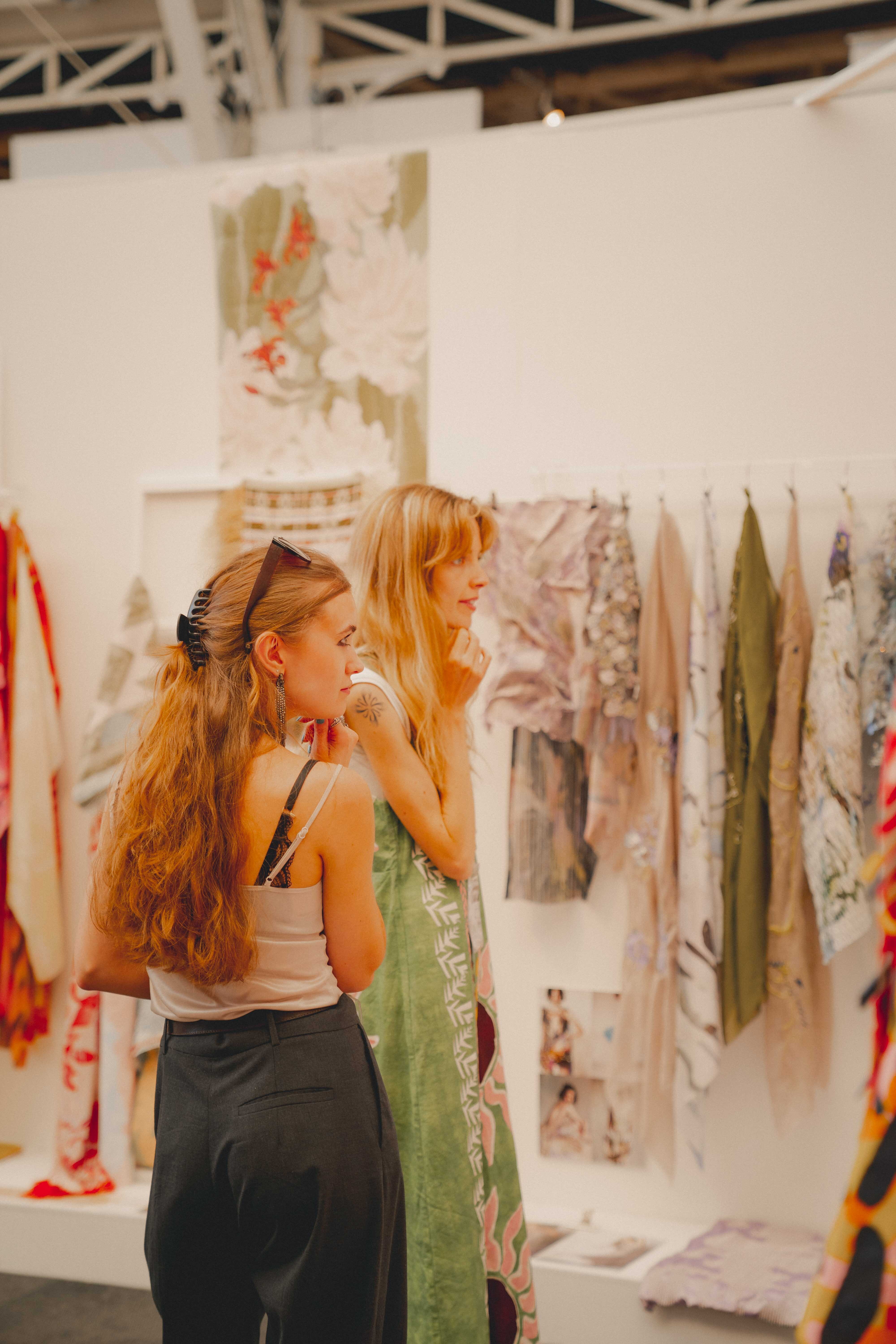 Two women browsing textiles, one in green dress, other in white top and black skirt, colourful textile displays visible in background.