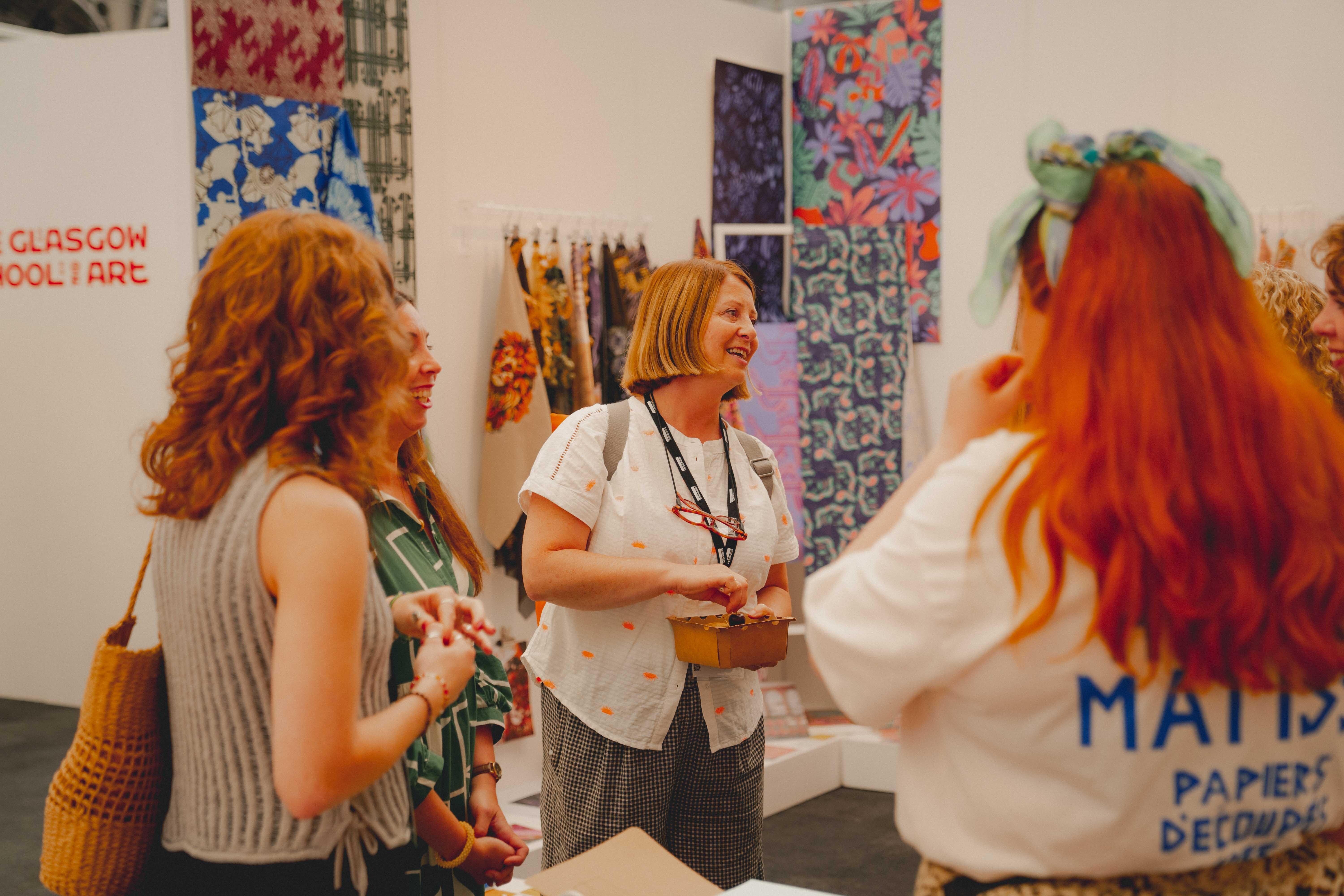 Three women with red hair conversing at art exhibition, colourful patterned textiles displayed on white wall behind them.