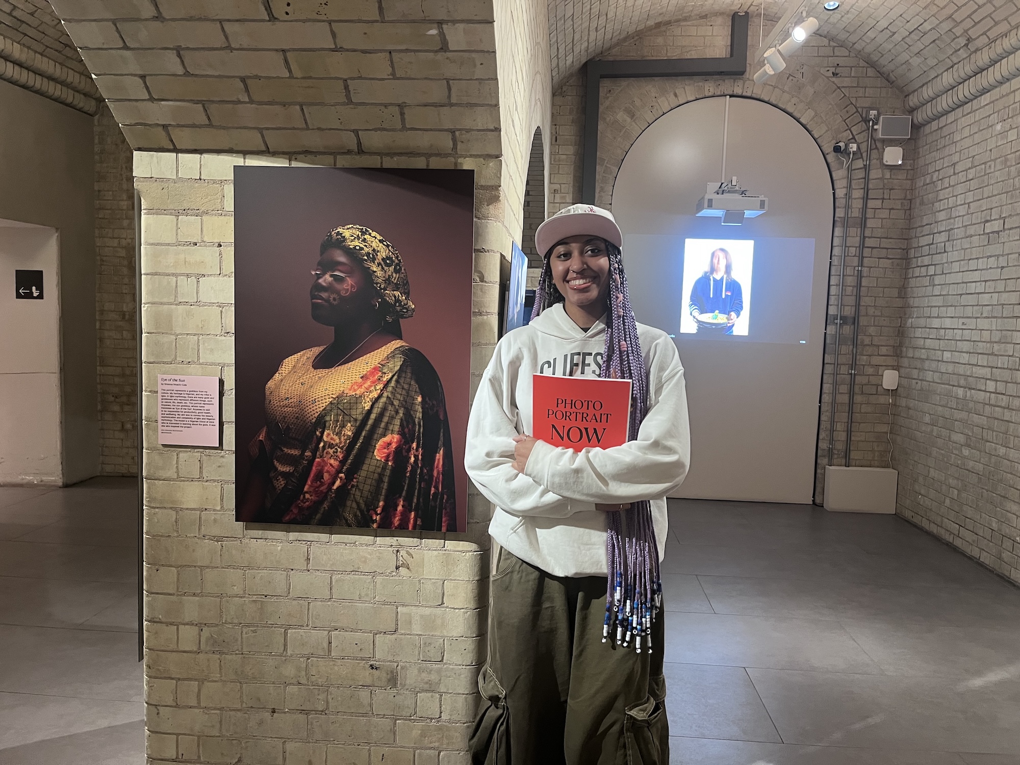 A person wearing a cap and long braids stands in a brick-walled gallery holding a red book, with a large portrait on the wall behind her and a projected image in the archway.