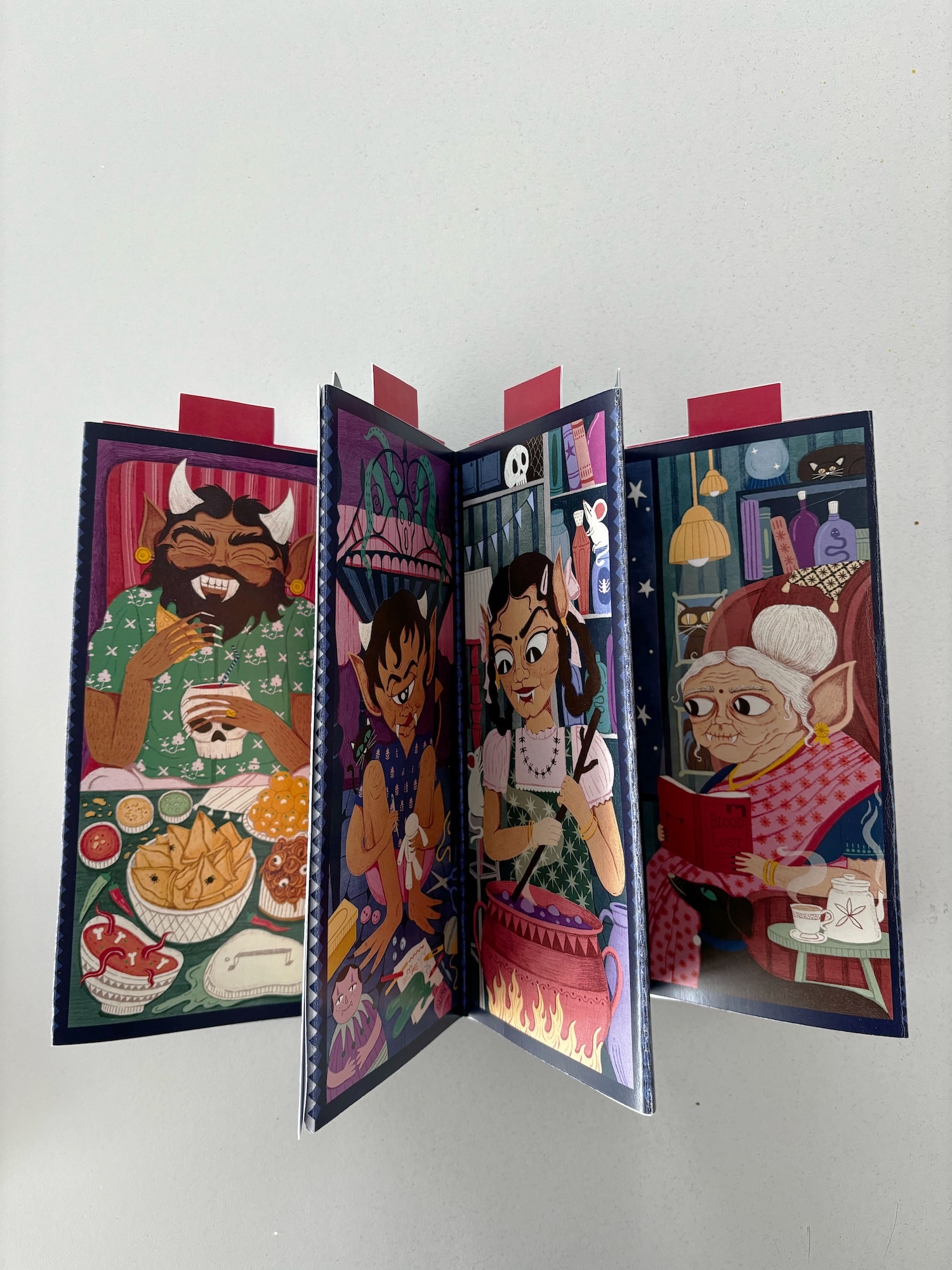 Four-panel folding illustrated book or card with colourful cartoon Indian folklore figures in different scenes, standing upright with red tabs visible.