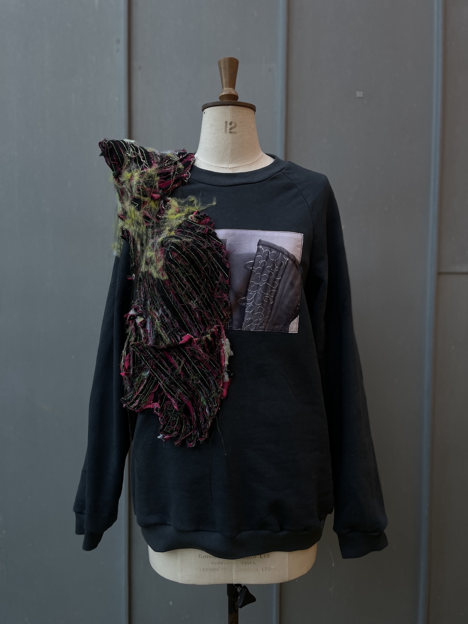 Black sweatshirt on mannequin with asymmetrical mixed-media design featuring printed graphics and textured fabric patches in dark colours with purple and green accents.