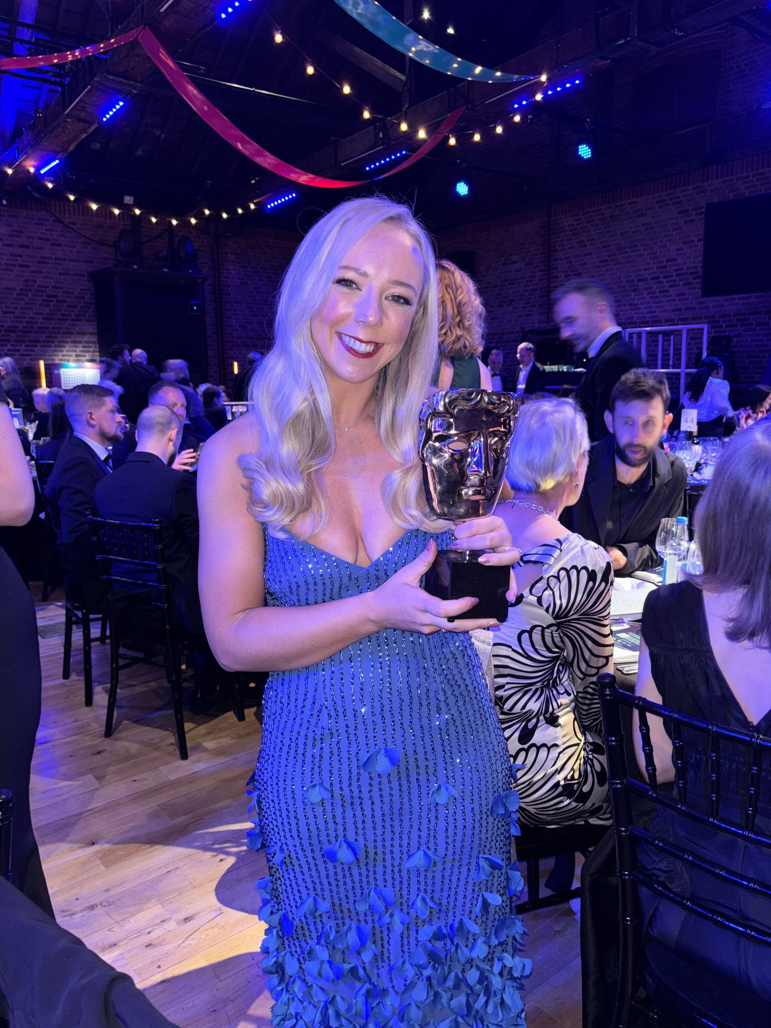 Make-up designer N Allan in blue sequined dress holding BAFTA Television Craft award at awards ceremony with fairy lights and guests in background.