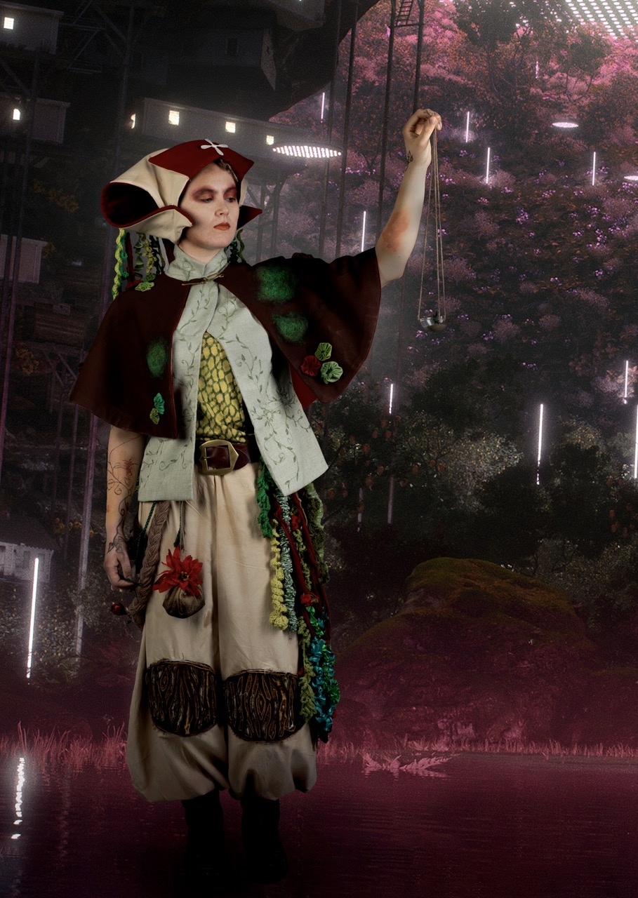 A person in an elaborate costume with a patterned headpiece and layered clothing stands with one arm raised, against a dark indoor backdrop with vertical light strips and pink trees.