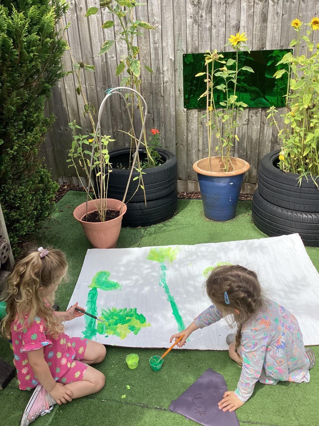 Two children sitting and painting on a large piece of paper in a garden area.