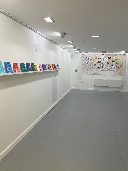 White gallery interior with colourful books on floating shelf, grey floor, recessed ceiling lights, and artwork on far wall.