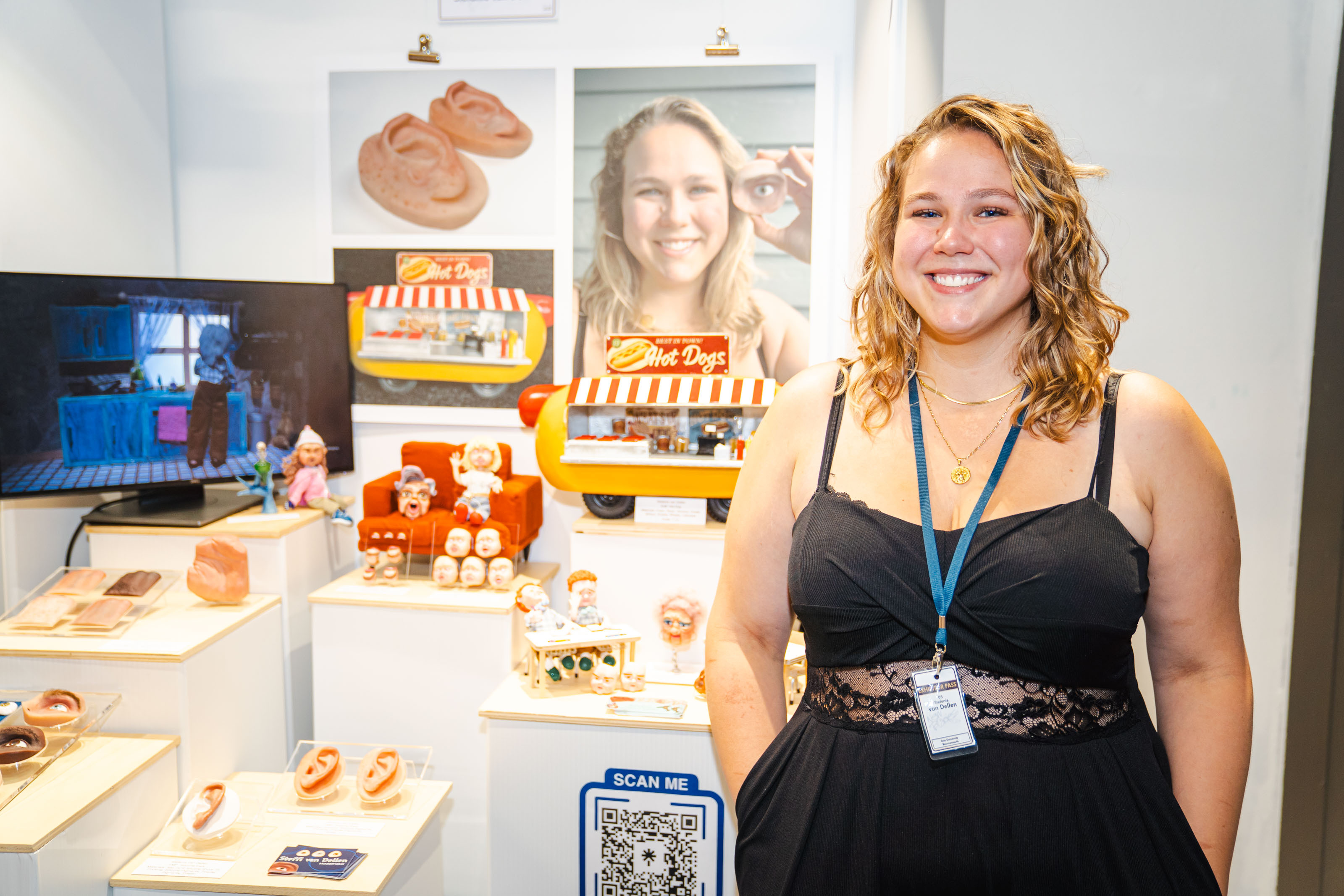 BA (Hons) Modelmaking graduate S van Dellen stands with bakery product models on display, branded packaging, and promotional materials on white table at New Blades Modelmaking expo.
