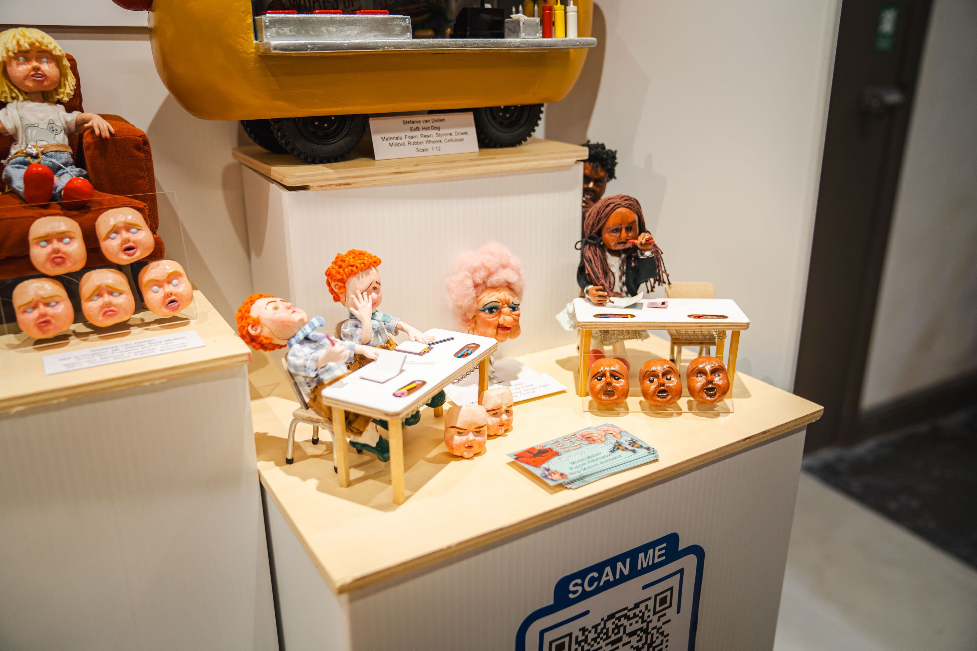 Museum display with miniature dolls, dollhouse furniture, and political figure masks on white pedestals with information plaques.