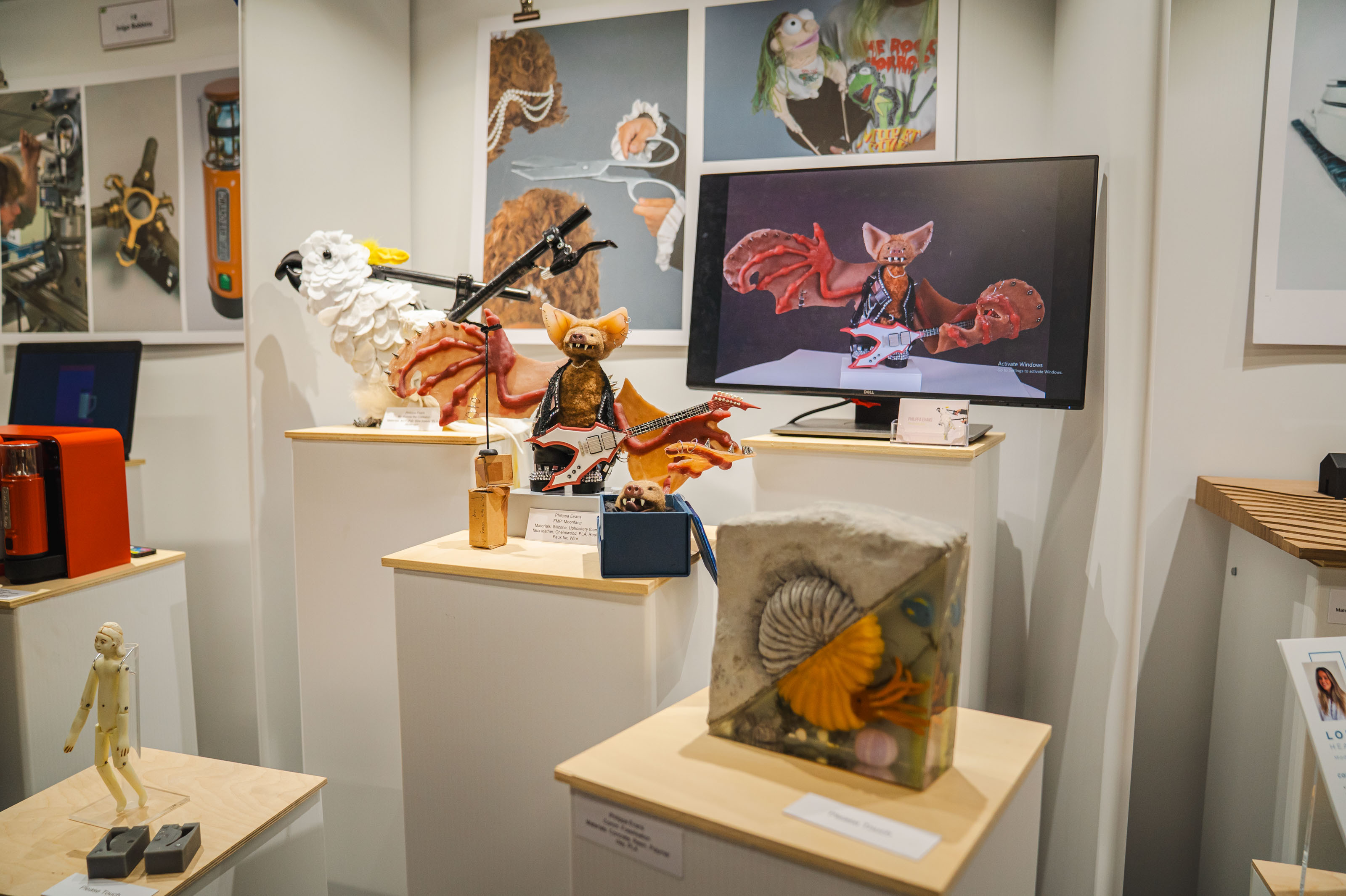 Display at New Blades Modelmaking expo with mixed media artworks on white walls and wooden shelves, featuring colourful illustrations and sculptural pieces.