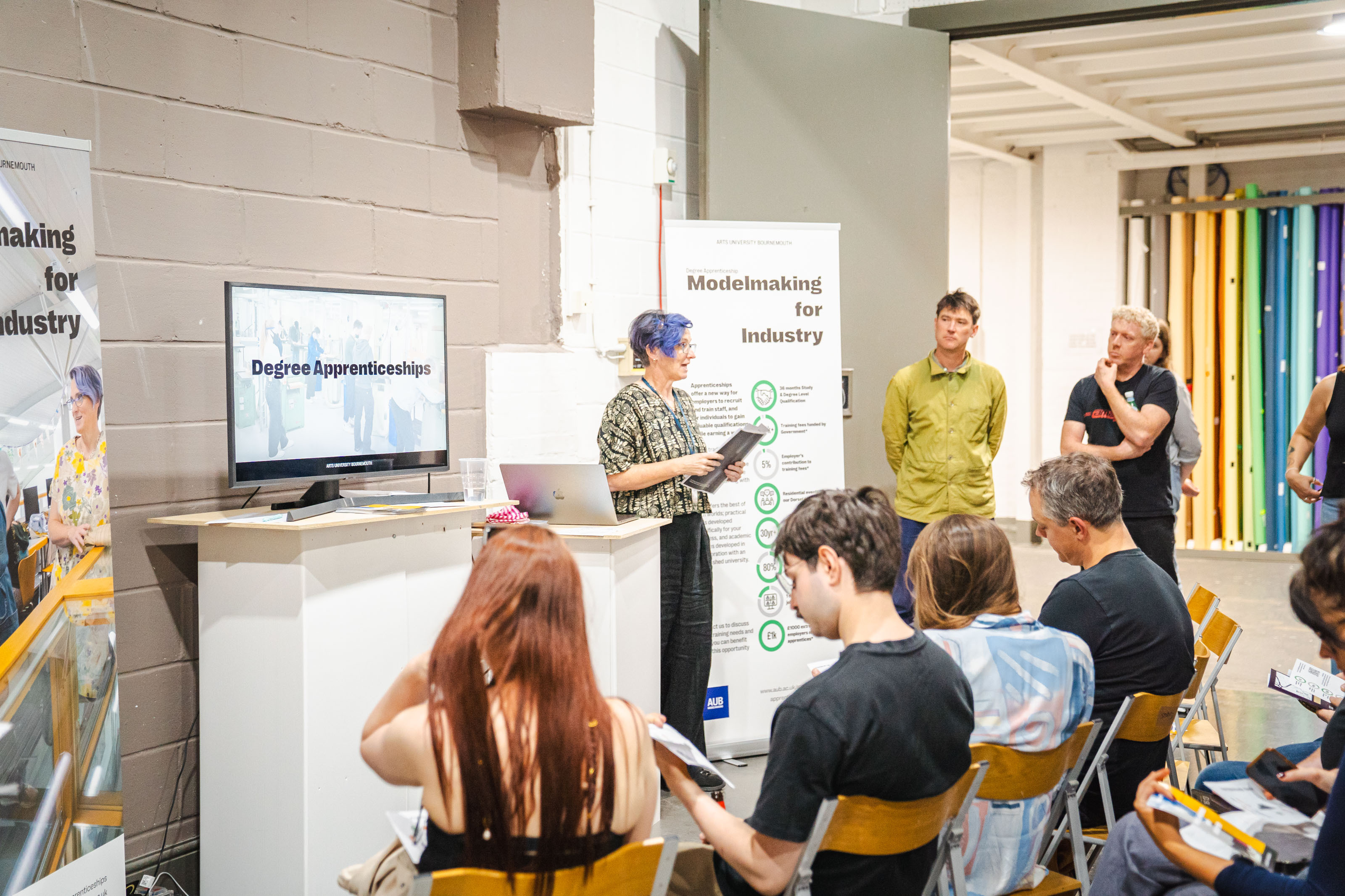 Dr C Holman presenting to seated audience in modern gallery space with white walls, colourful vertical strips, and "Modelmaking for Industry" signage visible.