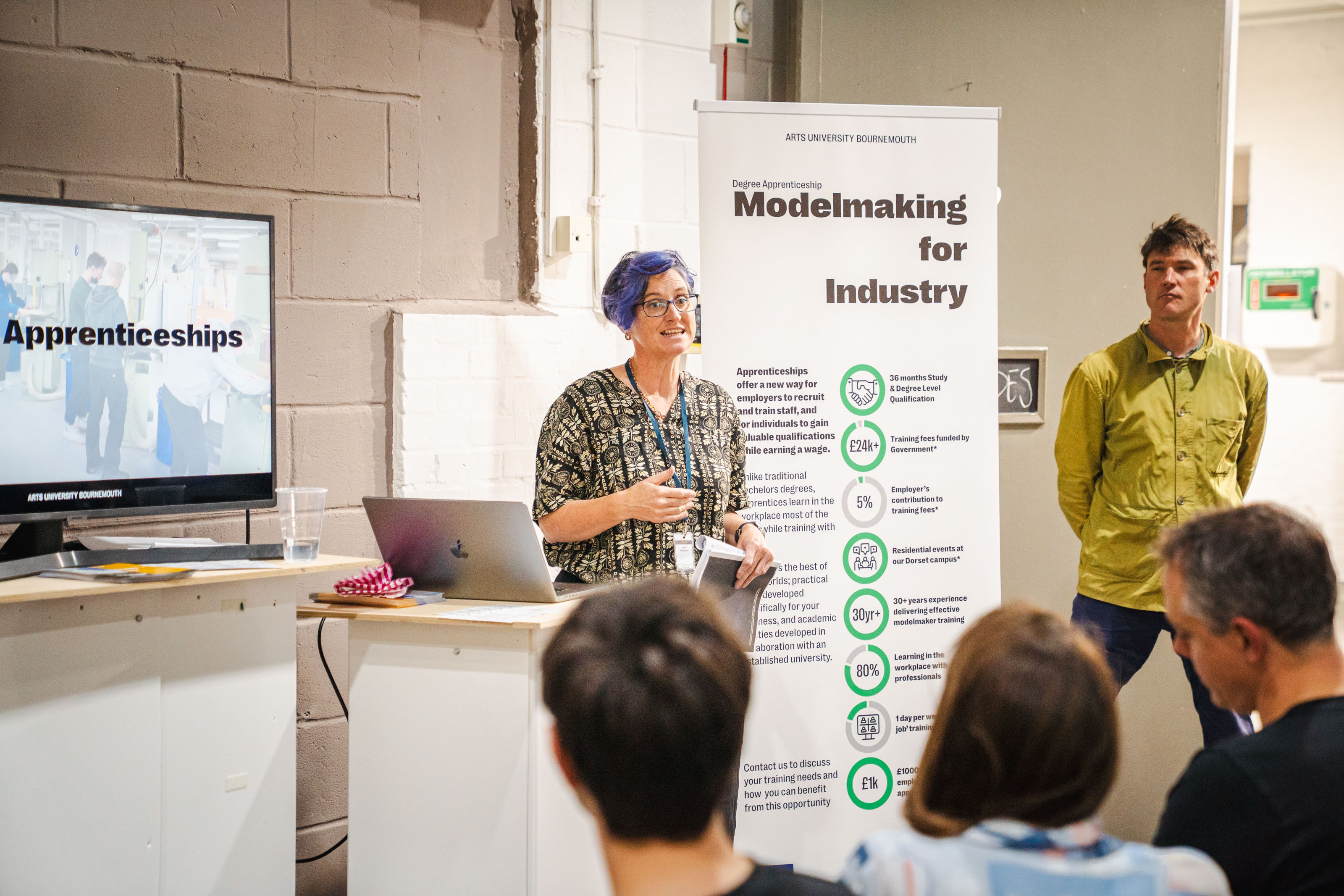Dr C Holman presenting to seated audience beside "Modelmaking for Industry" banner and "Apprenticeships" screen, with a person in green shirt standing nearby.