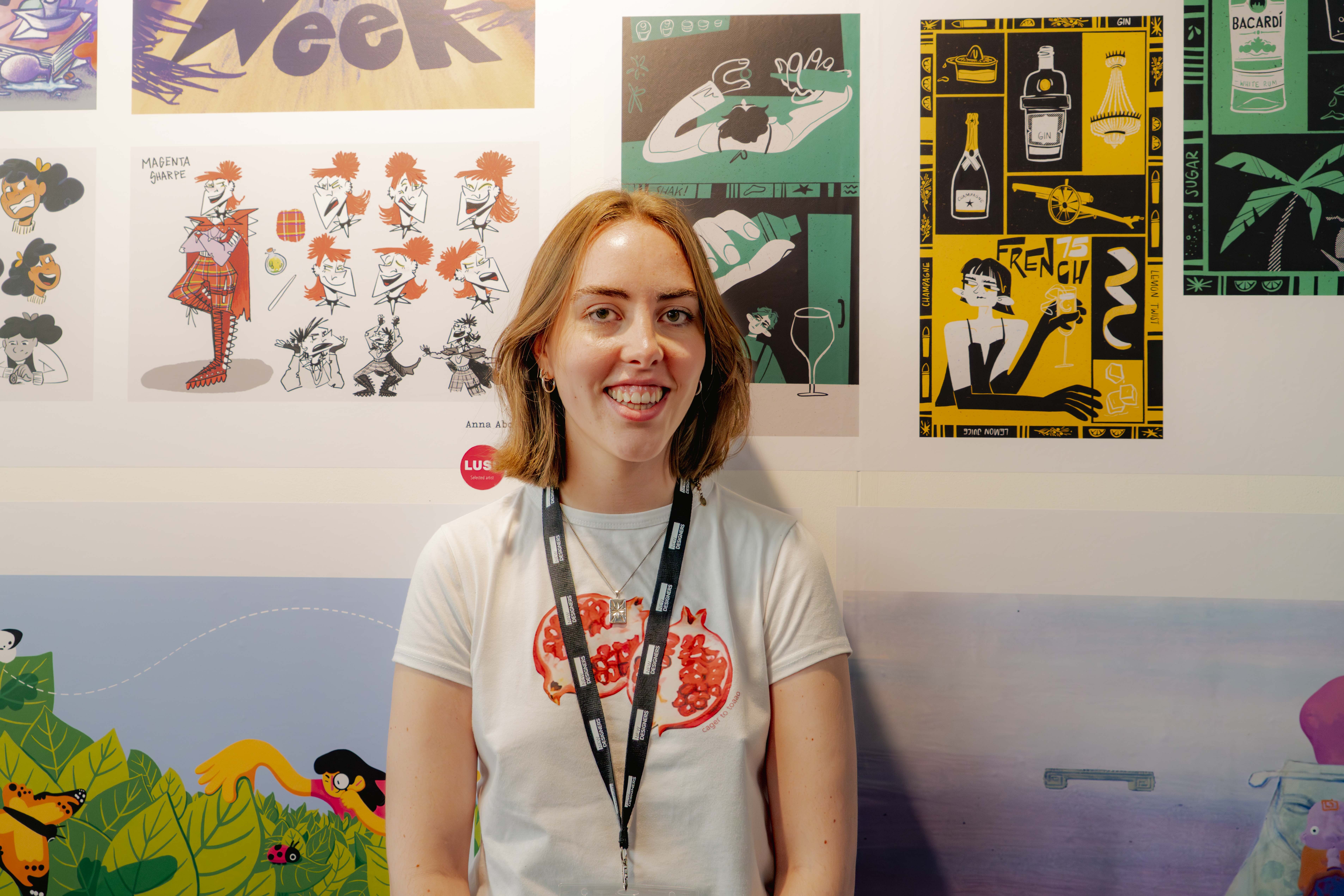 Young woman with shoulder-length brown hair wearing white t-shirt and black lanyard, smiling at camera against wall of colourful artwork.