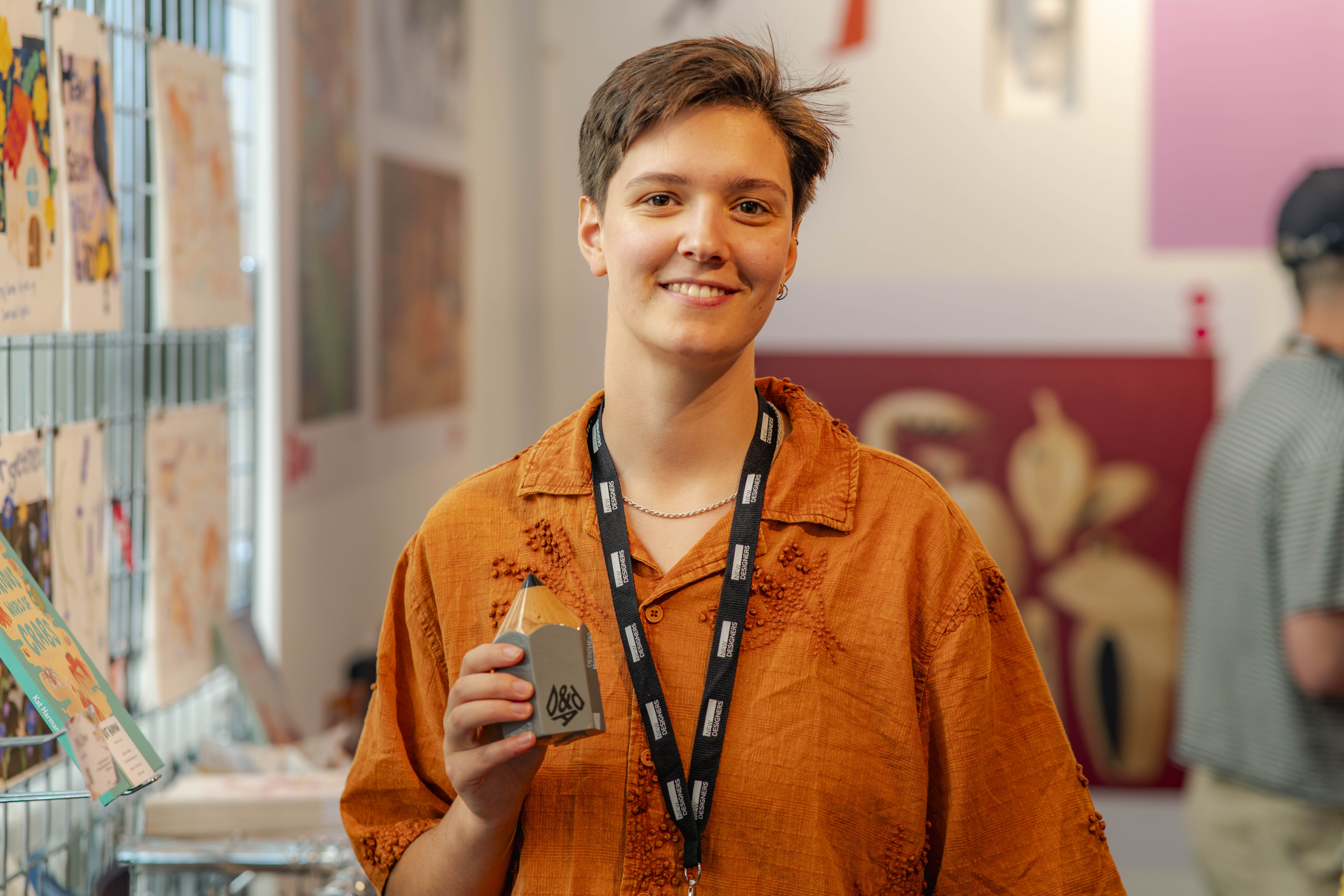 B O'Hara holding D&AD New Blood Graphite Pencil Award, smiling at stand at New Designers with colourful books behind.