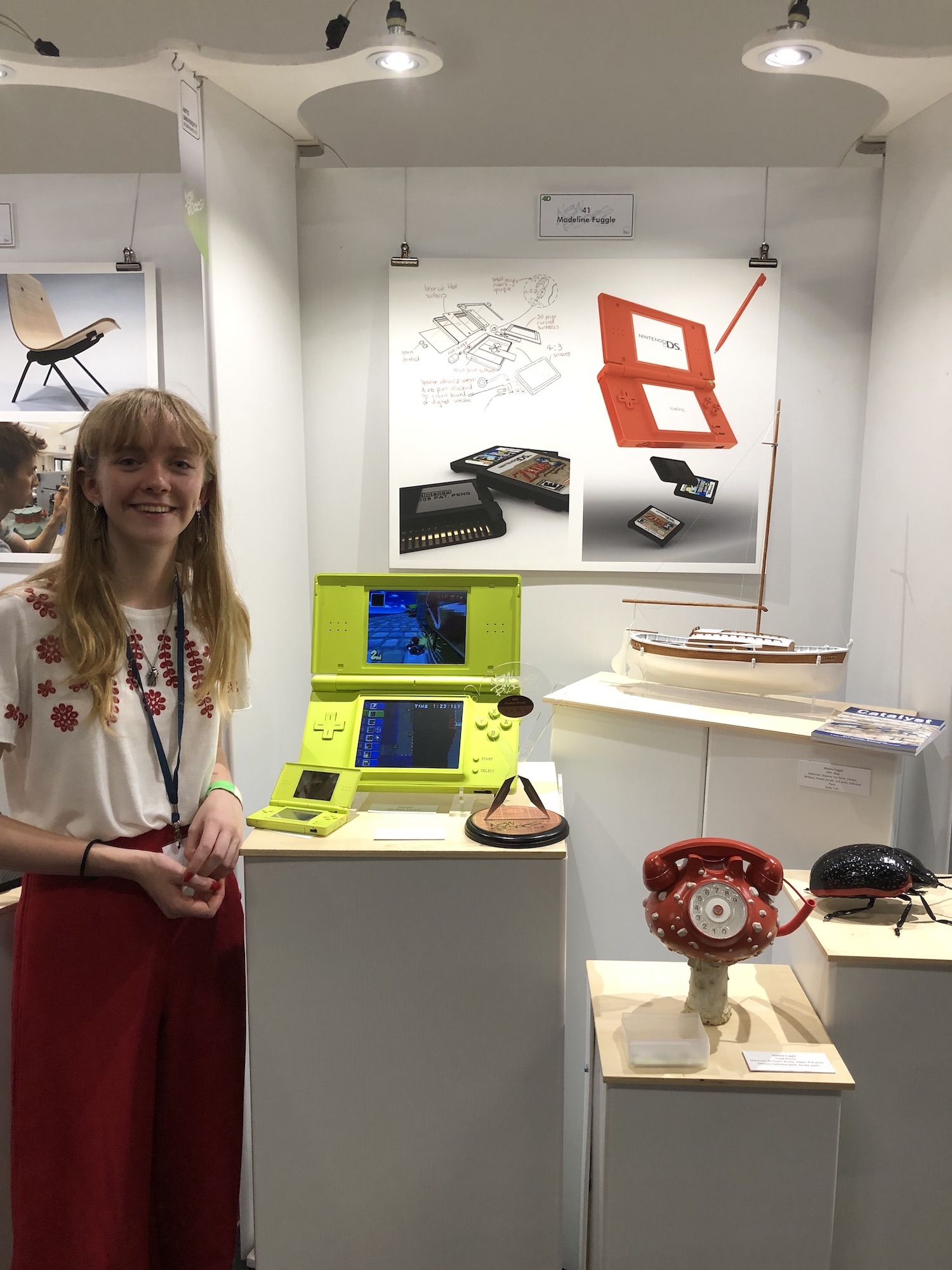 A BA (Hons) Modelmaking student at New Blades 2023 standing next to models of a Nintendo DS, a yacht, a telephone and a beetle.