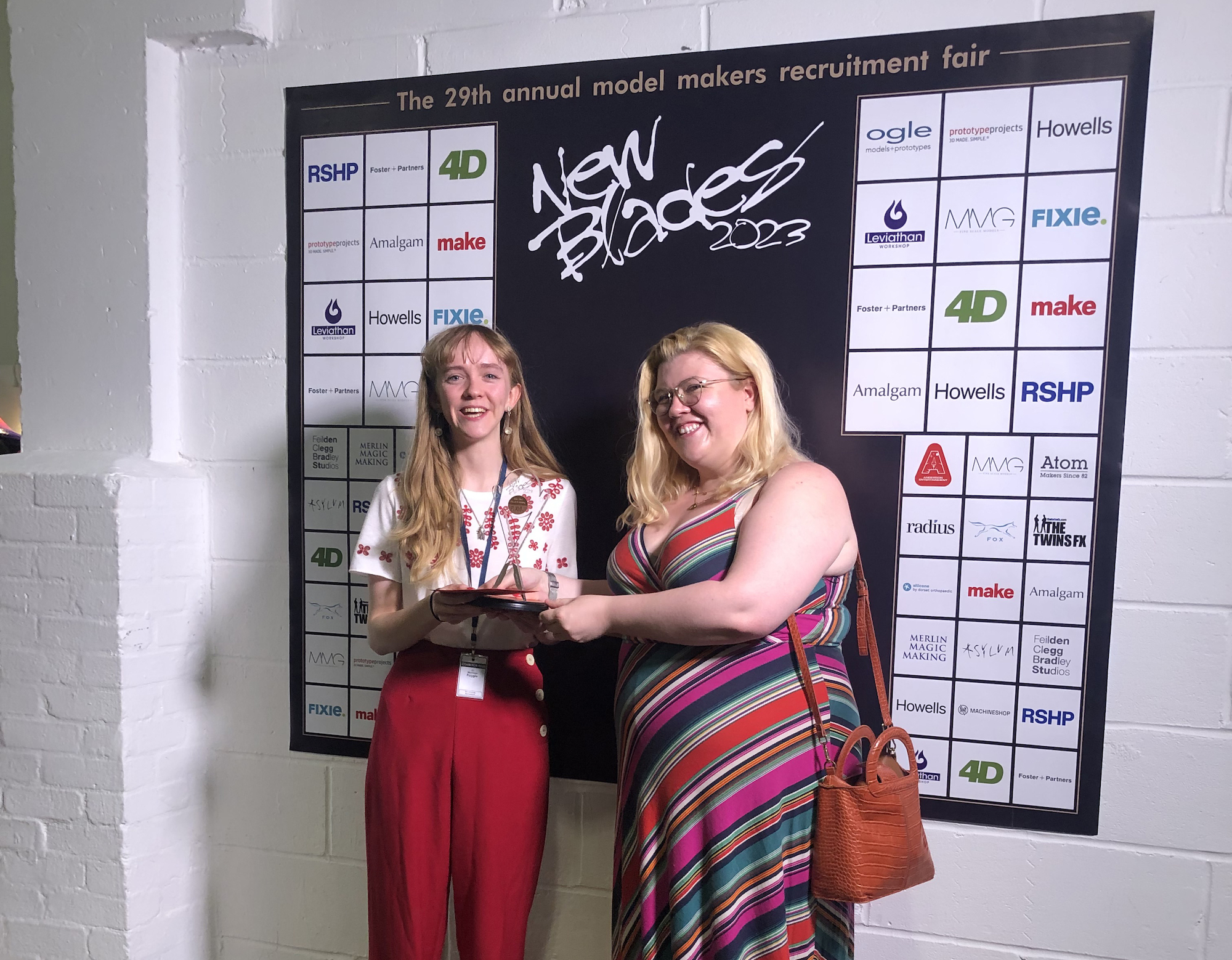 BA (Hons) Modelmaking student Maddie Fuggle poses in front of a large New Blades poster with different sponsor logos on it.