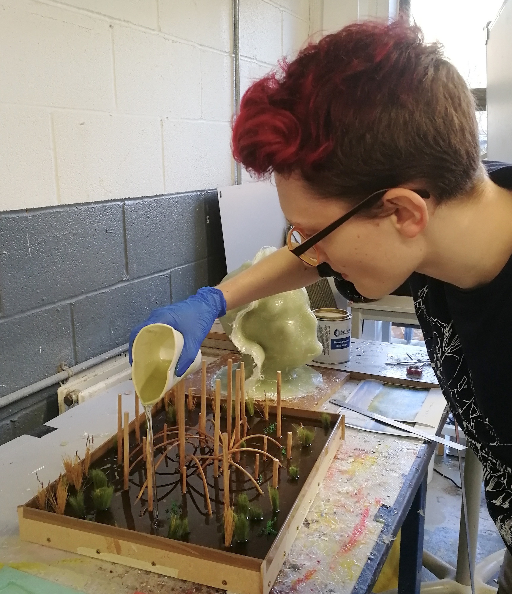 A Modelmaker pouring resin on a wooden structure diorama to mimic water, with mock plants.