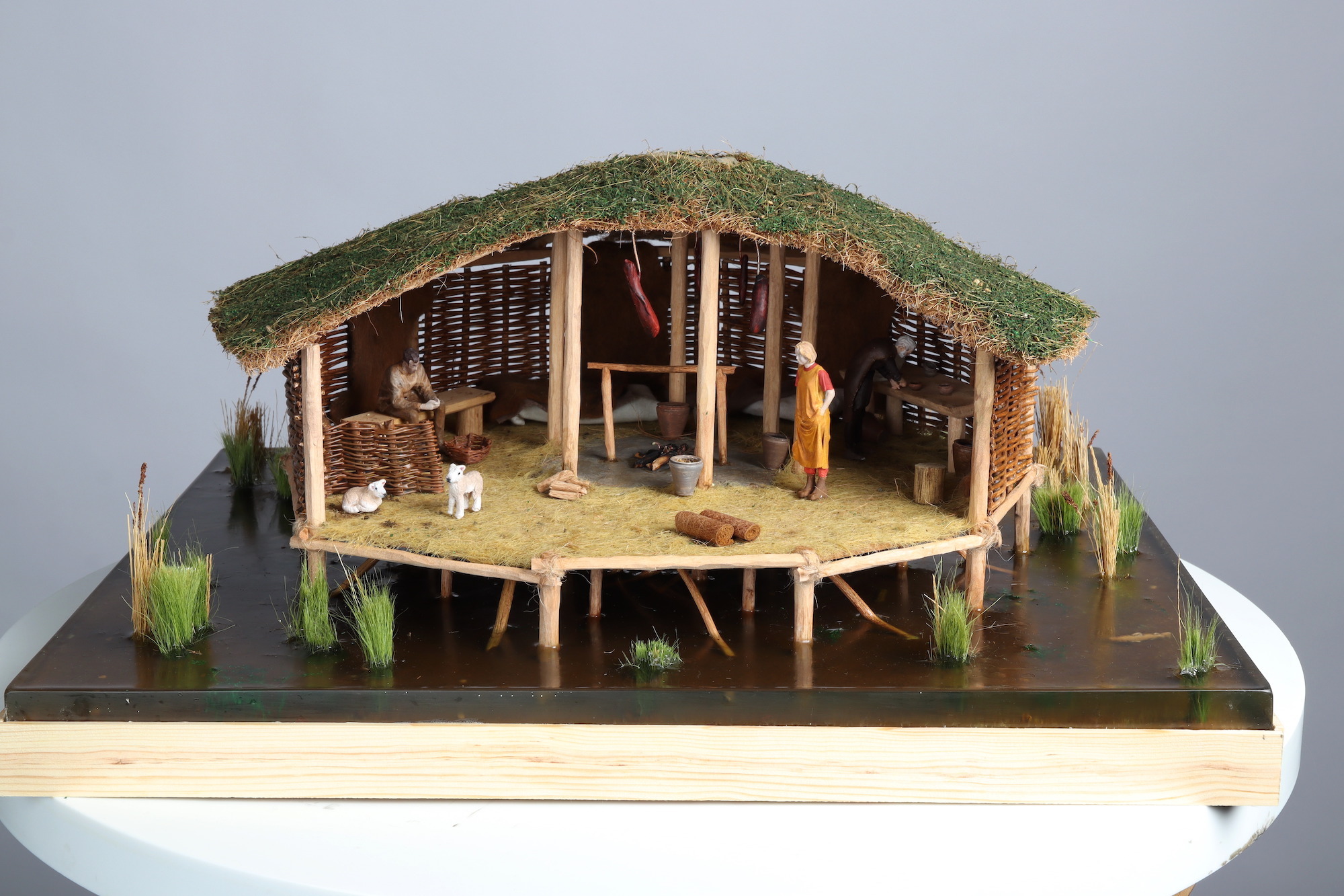 Small-scale cross-section model of a wicker roundhouse with a grass roof, people and creatures inside, white background.