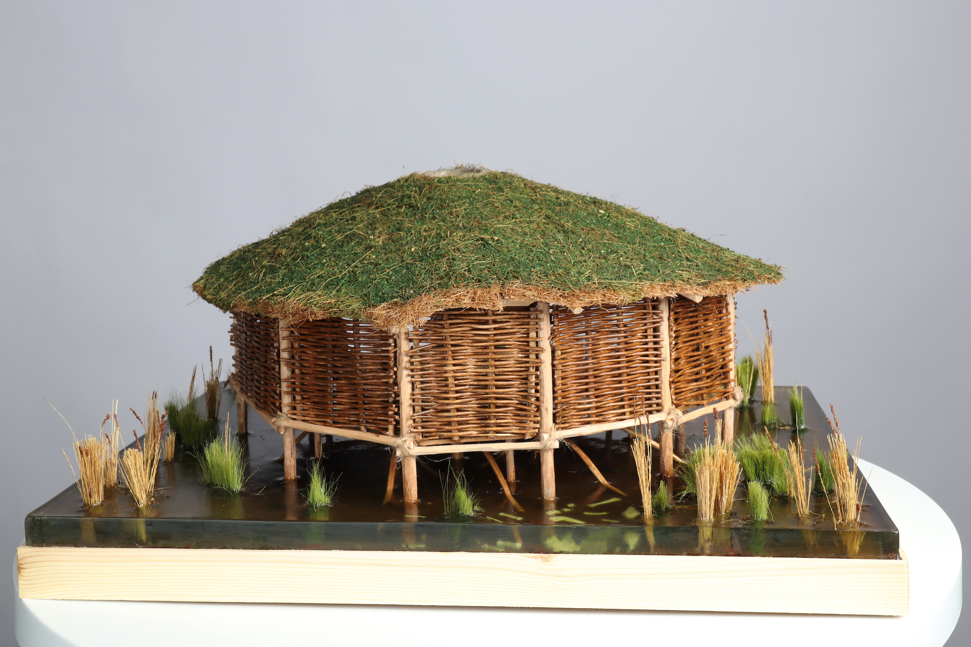 Small-scale cross-section model of a wicker roundhouse with a grass roof, back view, white background.