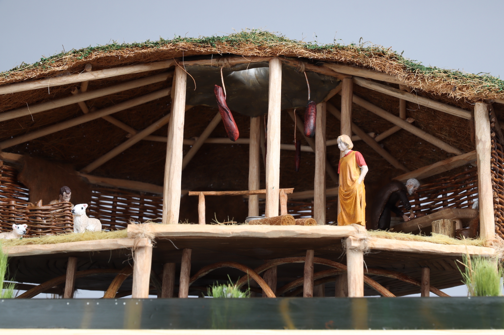 Small-scale cross-section model of a wicker roundhouse with a grass roof, people and creatures inside, white background, close-up.