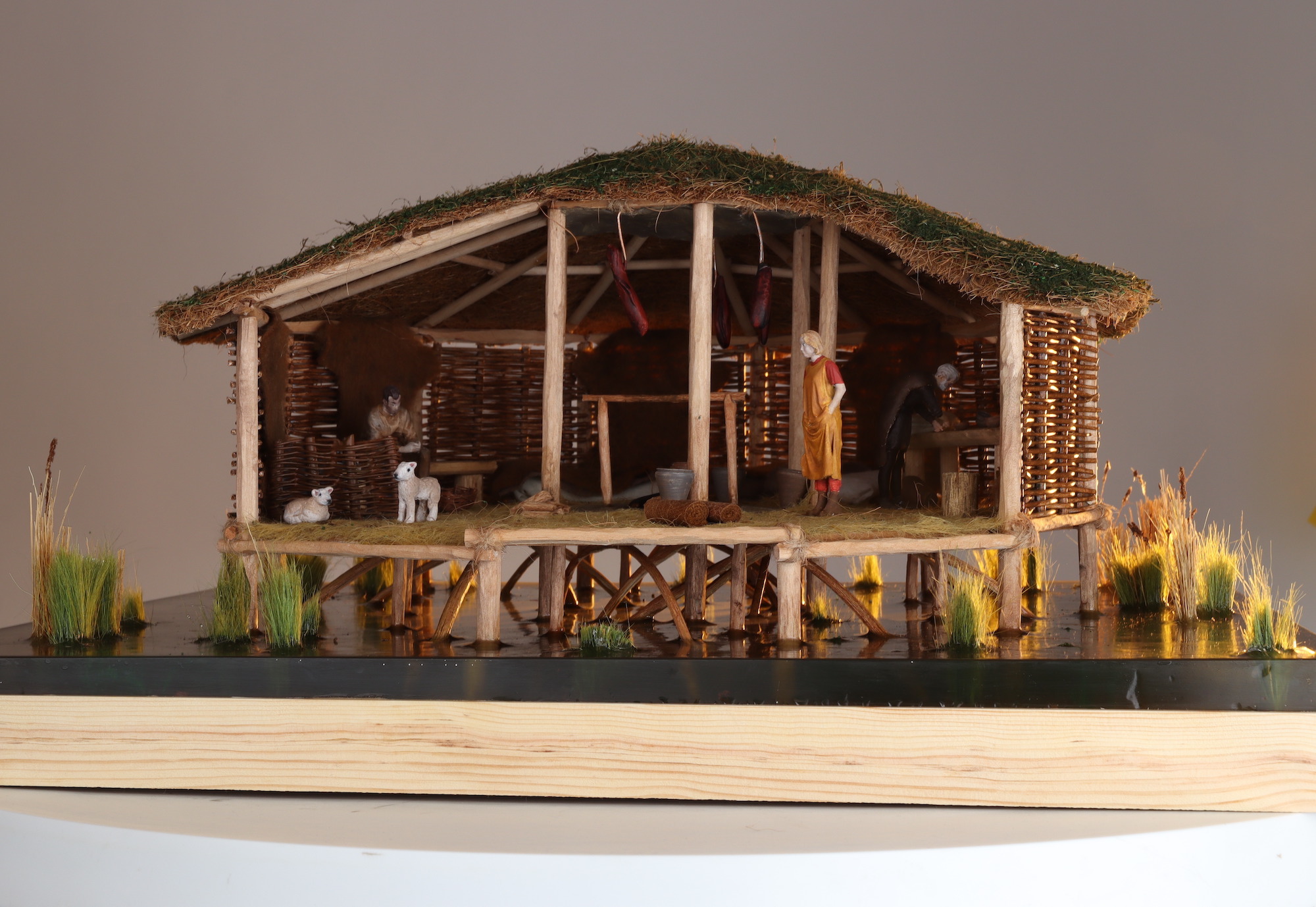 Small-scale cross-section model of a wicker roundhouse with a grass roof, people and creatures inside, white background.
