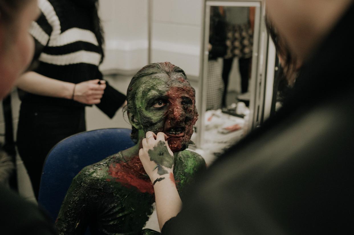 A person wearing makeup and prosthetics to look like a monster has more makeup applied.