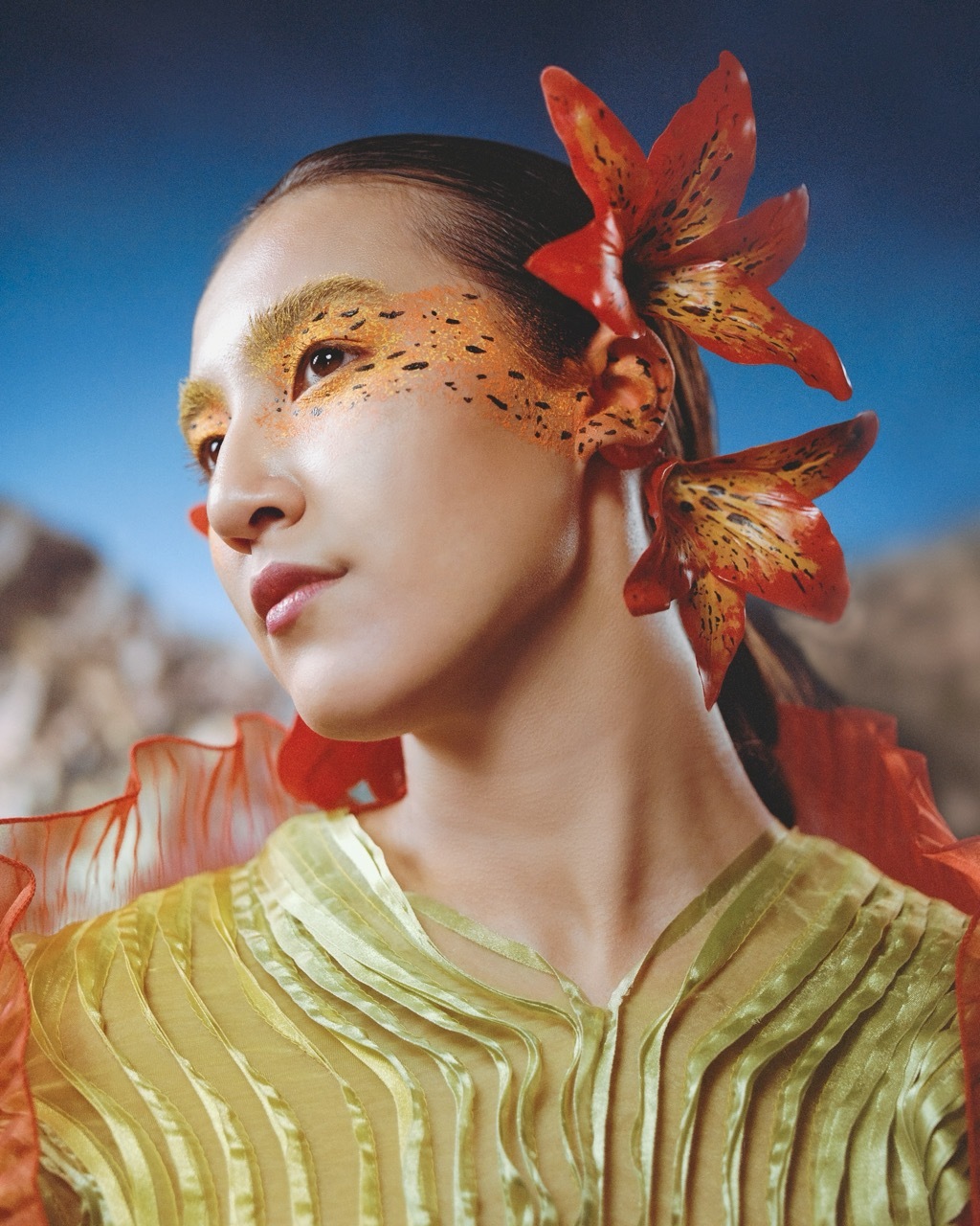 Model wearing red lilies with orange and yellow make-up, blurred background of Durdle Door in Dorset and a blue sky gradient.