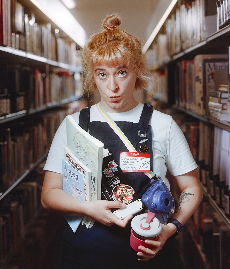 Portrait of a person pouting at the camera between two bookshelves, a bundle of objects in their arms.