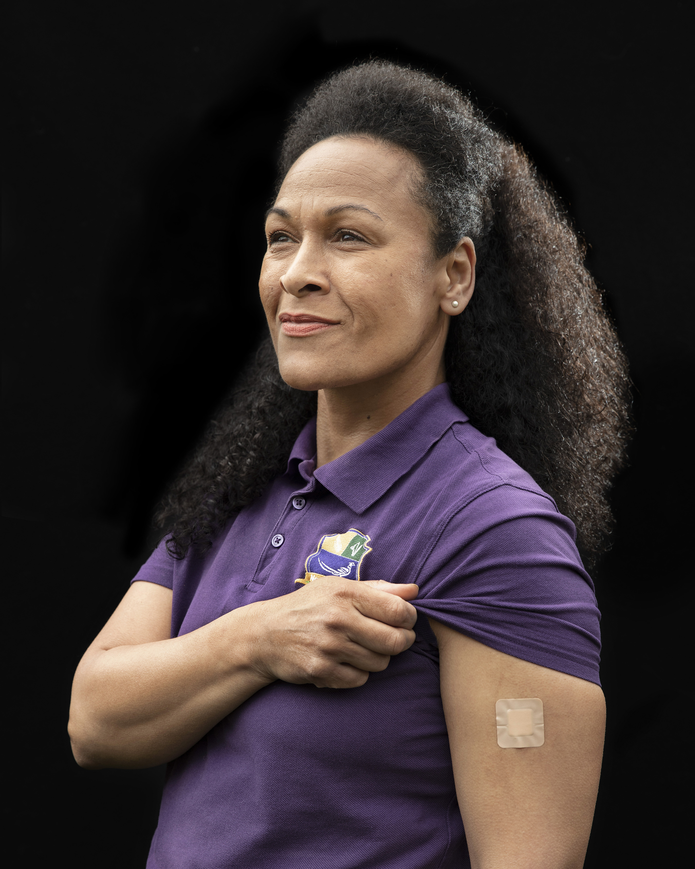 A woman looks off to the left and lifts their sleeve up to reveal a small plaster on their arm.