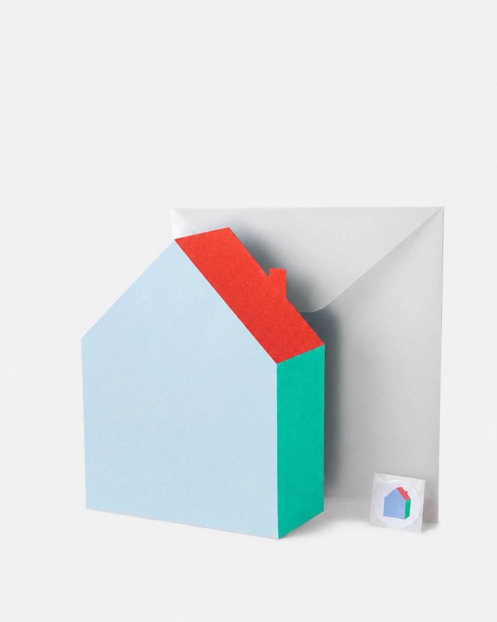 A minimalist paper house model with a light blue front, red roof and green side stands against a folded grey paper backdrop, with a small icon tile beside it.