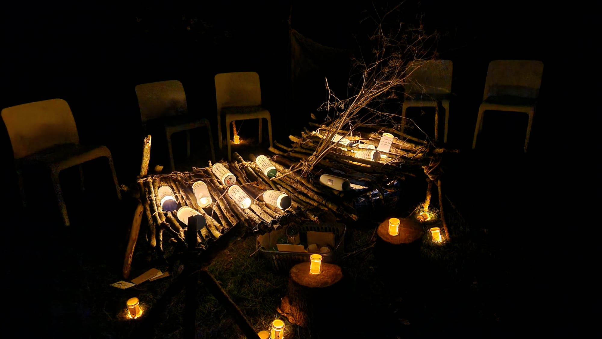 A small outdoor candlelit arrangement of wooden sticks and branches on a dark ground, with several chairs placed around it.
