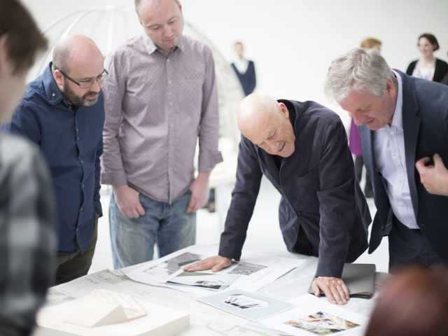 Four people stand by a table and look over a series of documents