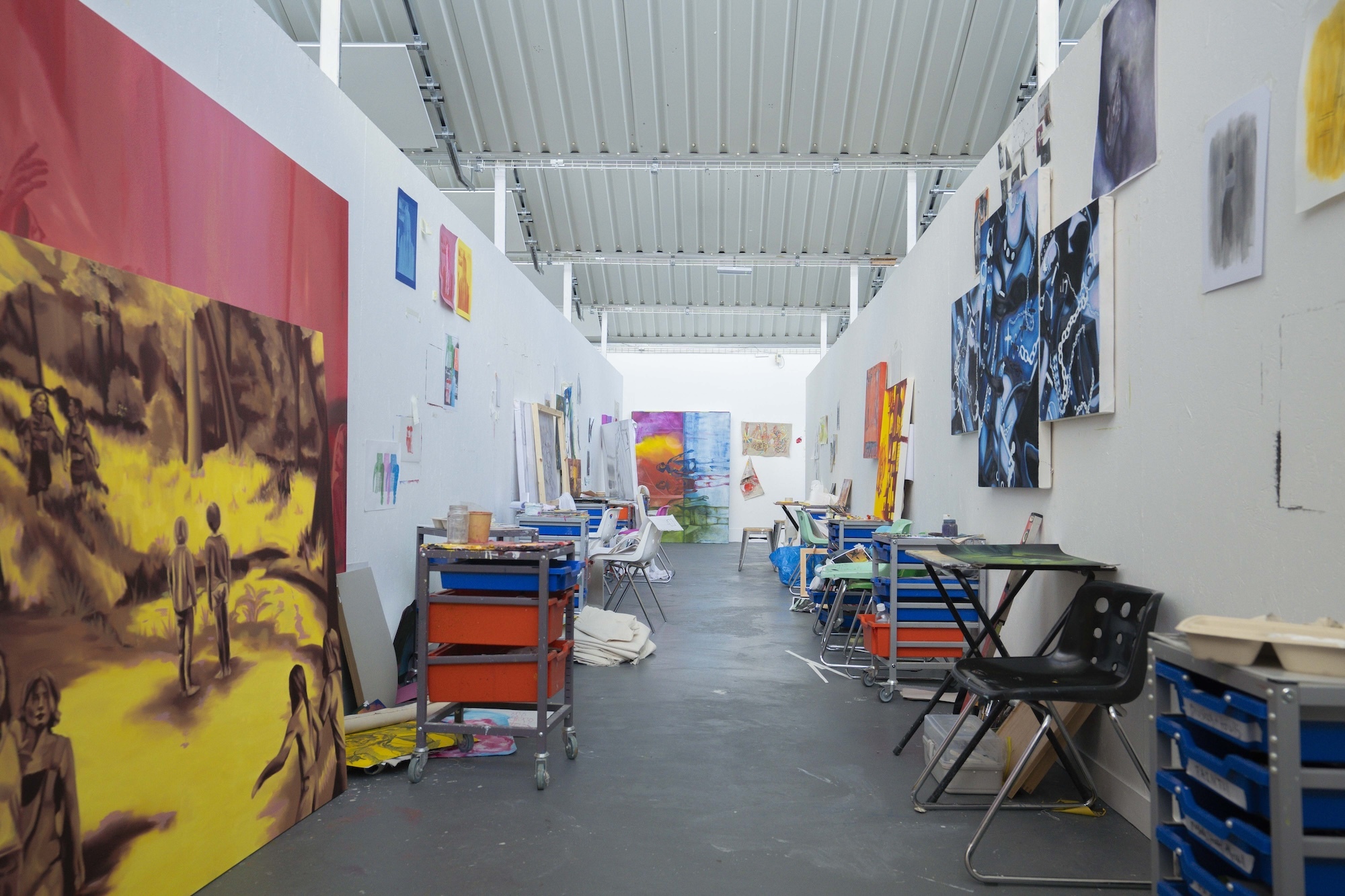 Artworks on display either side of a gallery studio space, with trolleys and drawers of artist supplies in the gangway.
