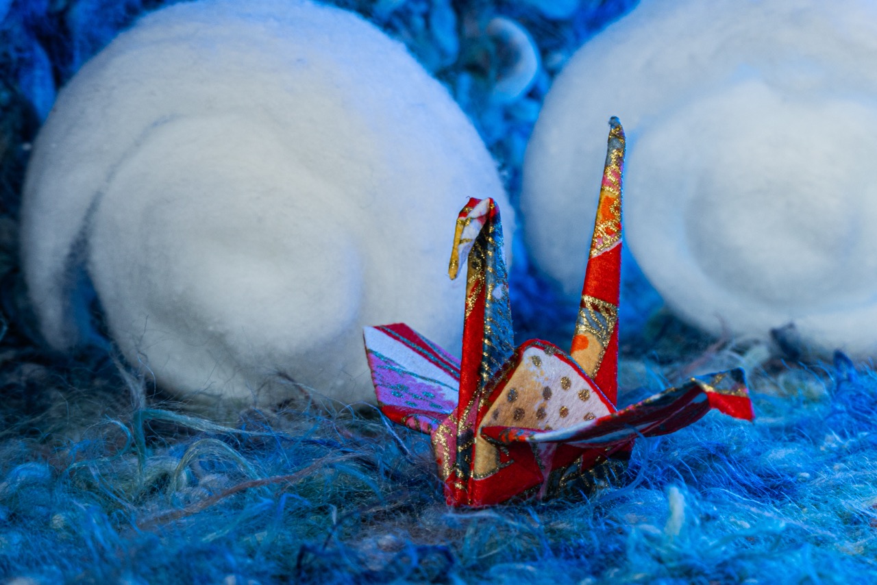 An origami crane made from patterned red paper on a blue fluffy surface, with two white wool balls behind it.