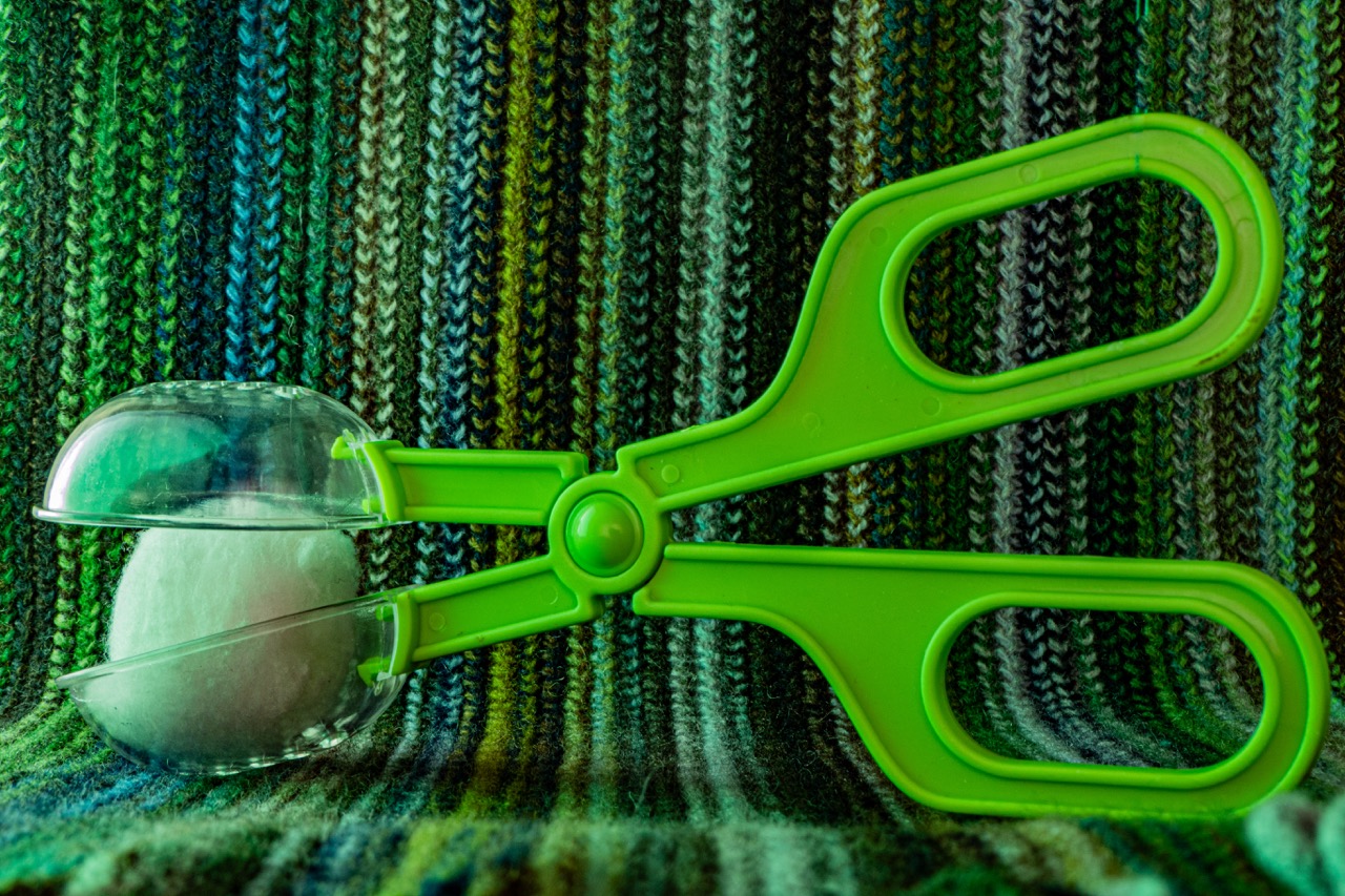 Green plastic scissors with a transparent egg-shaped cutter attached, placed on a multicoloured knitted fabric background.